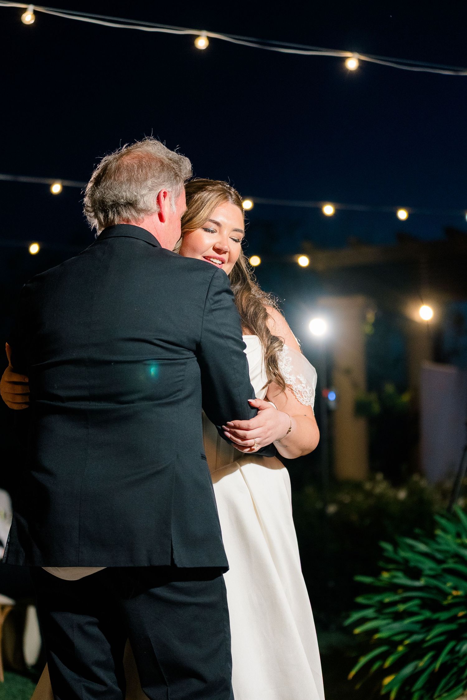 Emotional bride dancing with father under string lights at outdoor evening wedding reception.