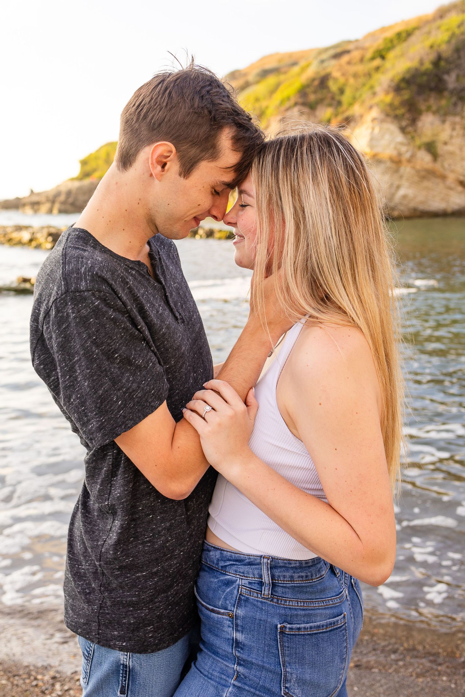 A couple sharing an intimate moment on a beach with rocky cliffs and waves in the background.