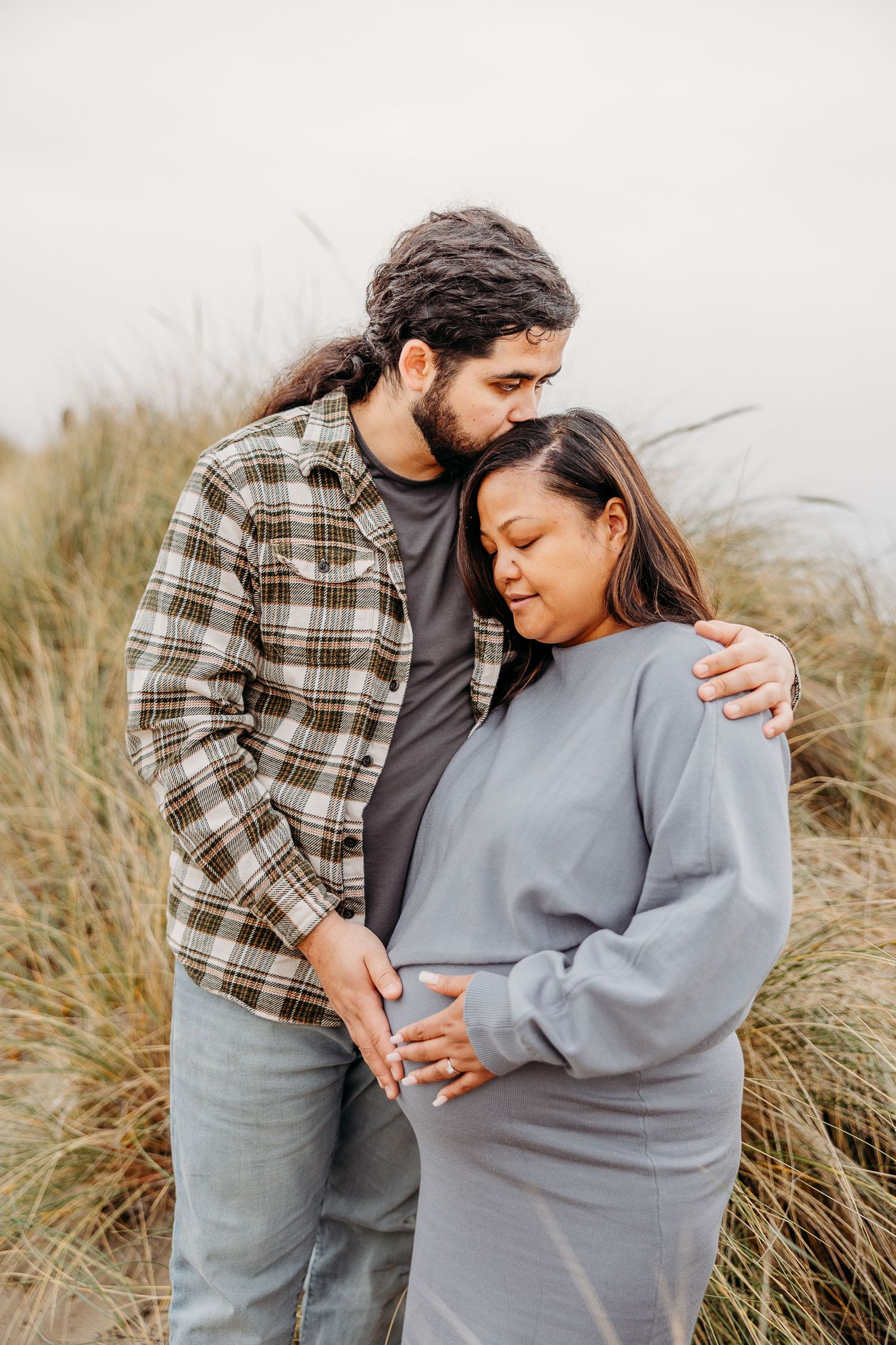 A couple shares an intimate moment together in casual sweaters among tall beach grass during a cloudy day.