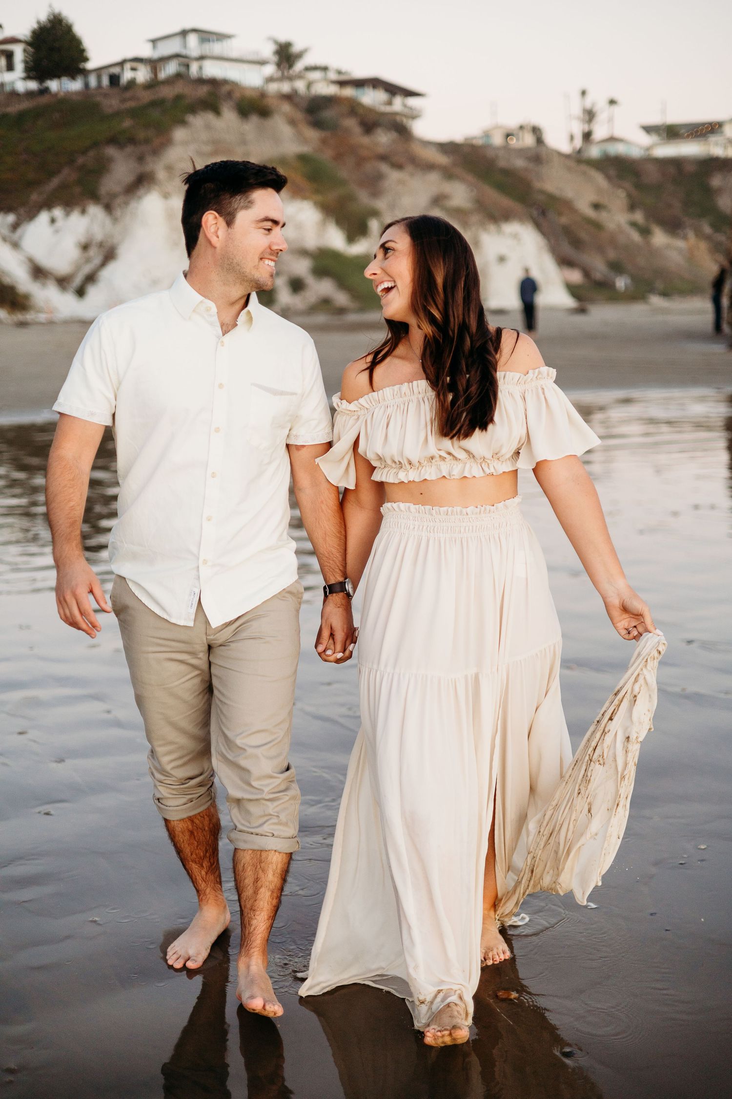 A couple walking hand in hand on wet sand beach wearing coordinated white and beige outfits.