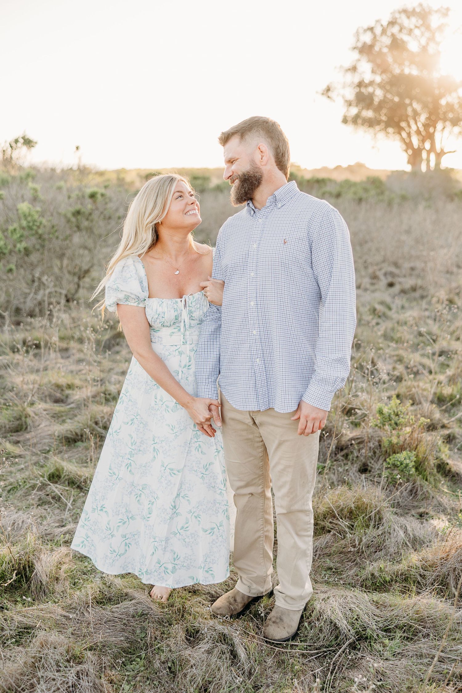 A romantic couple in coordinating blue and white outfits pose together on a grassy hillside at sunset.