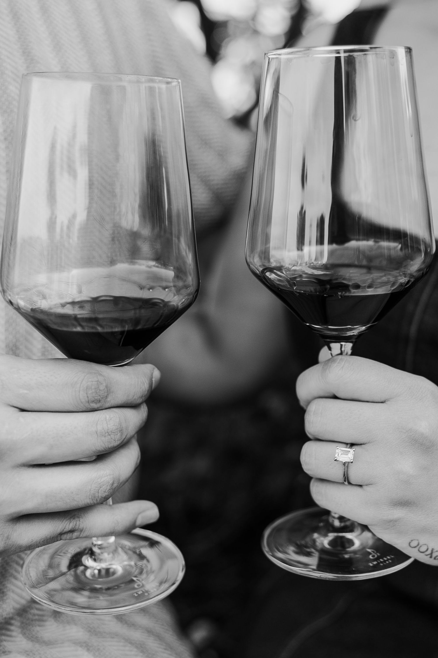 Two hands holding red wine glasses in a black and white photo with an engagement ring visible.