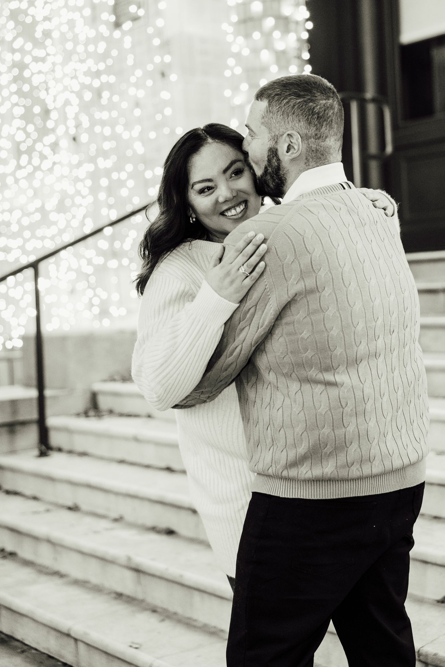 A romantic black and white photo of a couple embracing on steps with twinkling lights in the background.