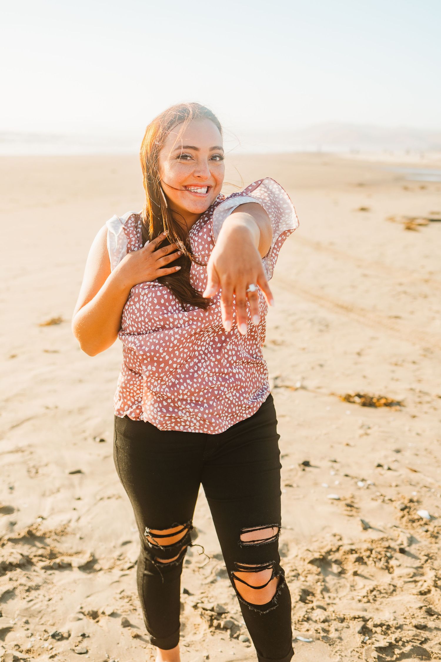 Woman in floral top and distressed jeans shows off engagement ring on beach at sunset.