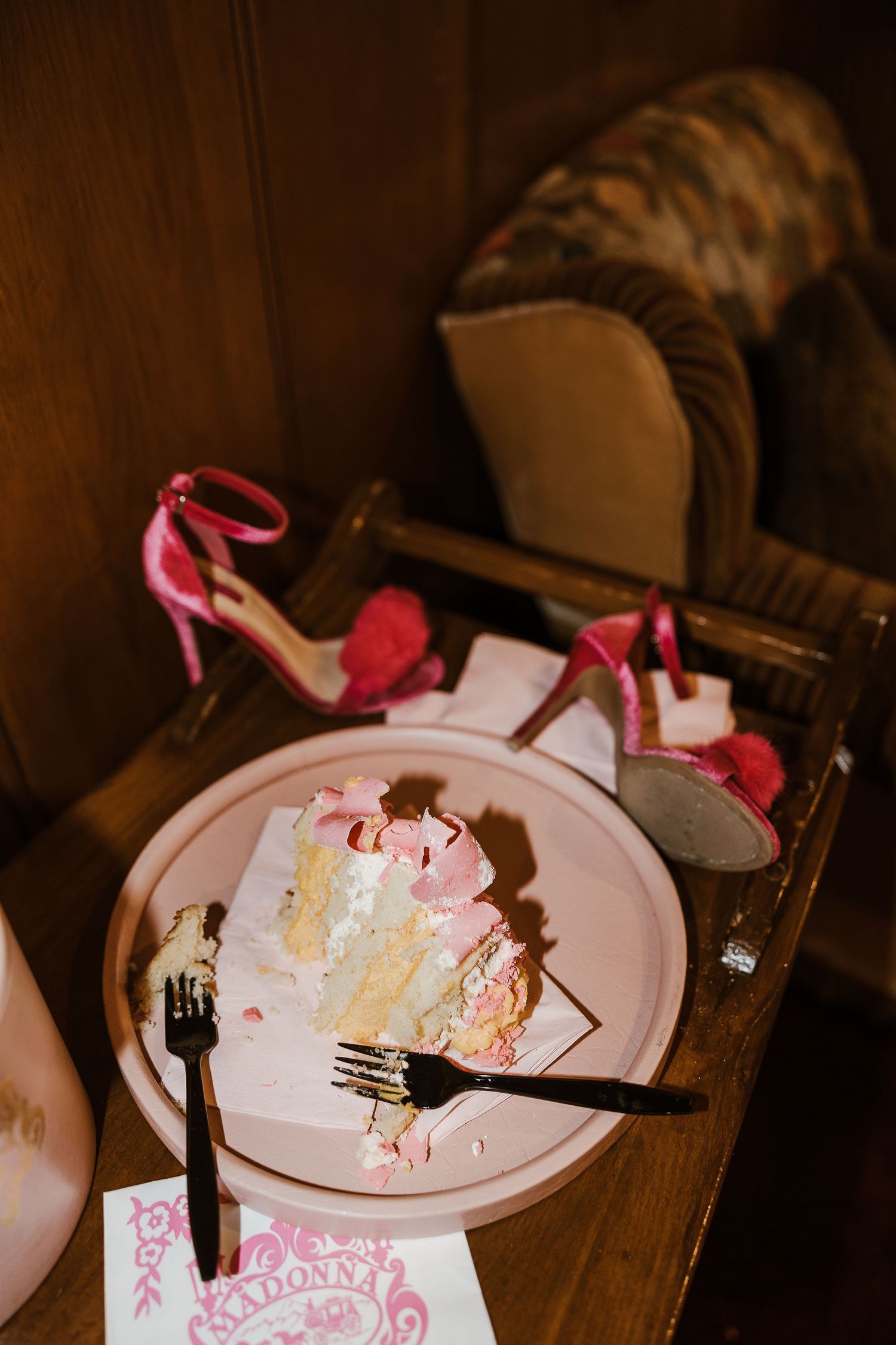 A half-eaten slice of pink-frosted cake on a plate with high heel shoes visible in the background.