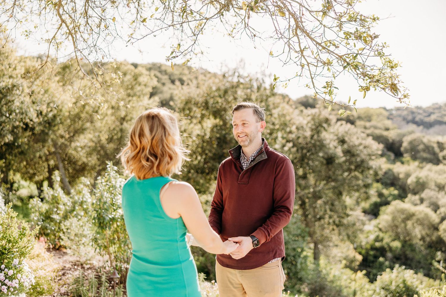 Two people stand together outdoors on a hillside path during golden hour with trees and mountains in background.