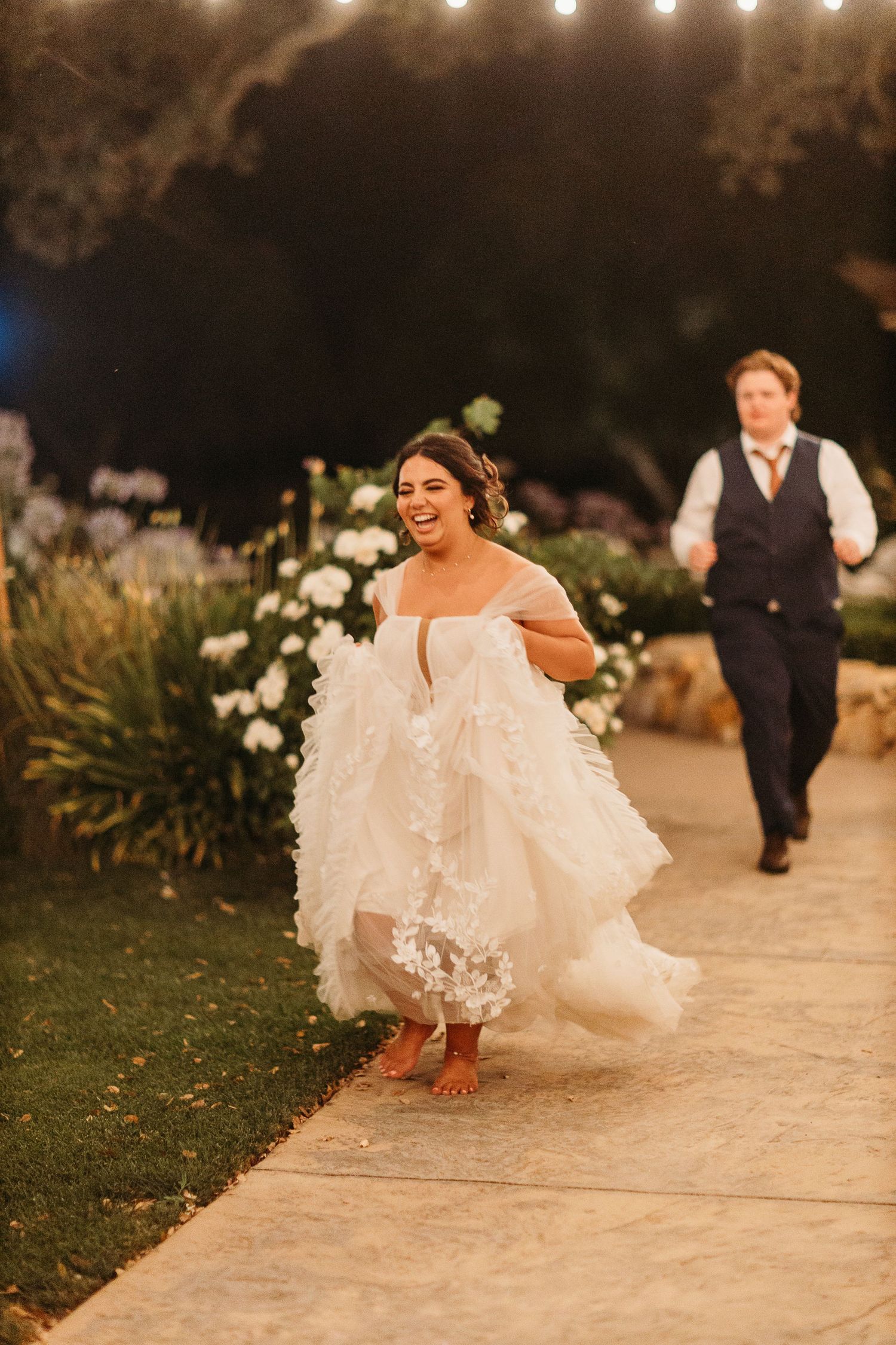 Joyful bride in white dress running along garden path during evening celebration.