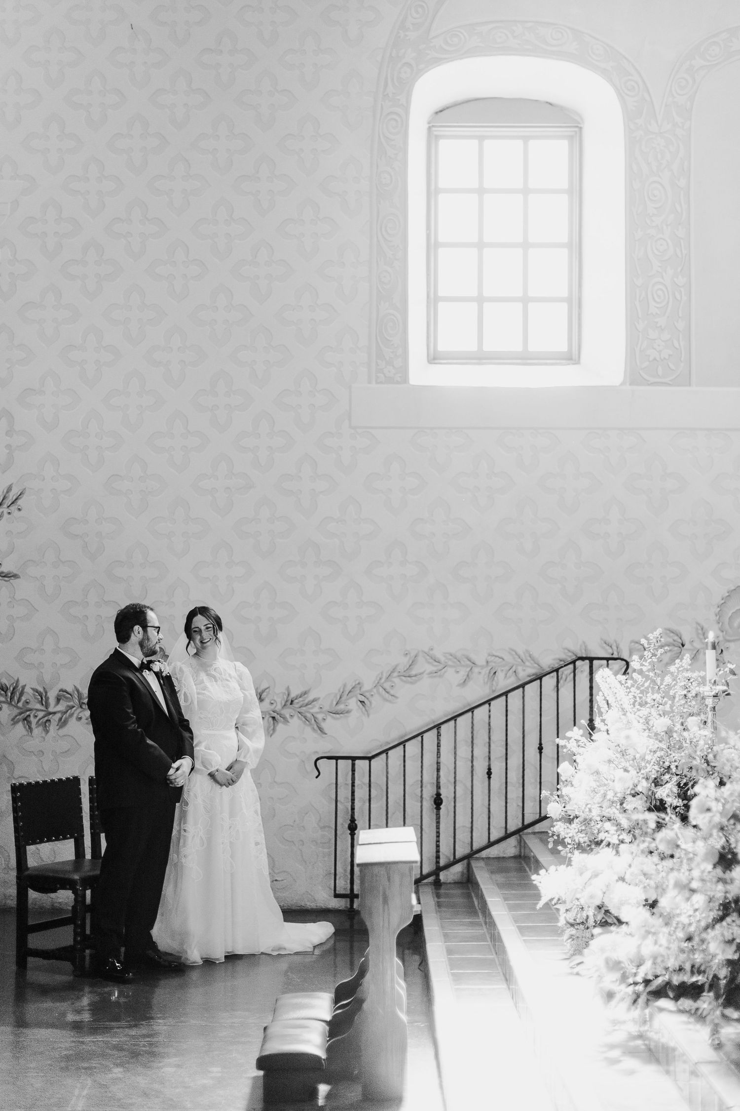 Black and white photo of a couple in formal attire standing by an arched window and floral arrangements during catholic ceremony in SLO.