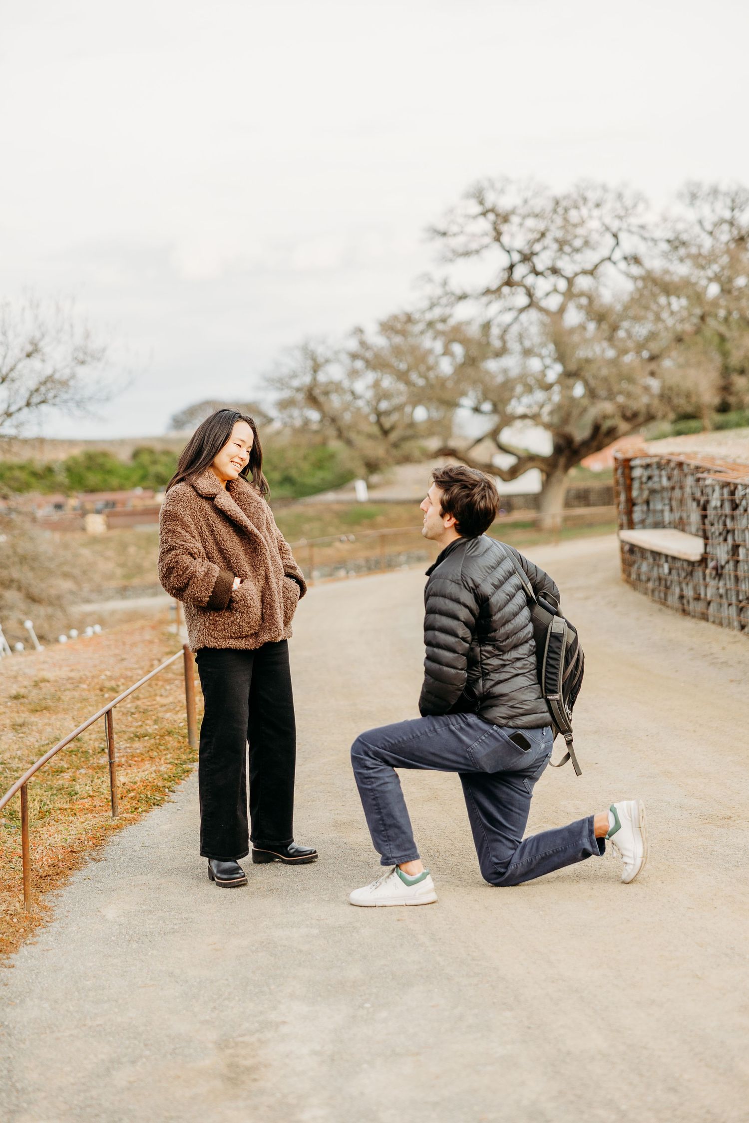 Two people in casual winter wear kneel on a path while taking photos outdoors on a cloudy day.