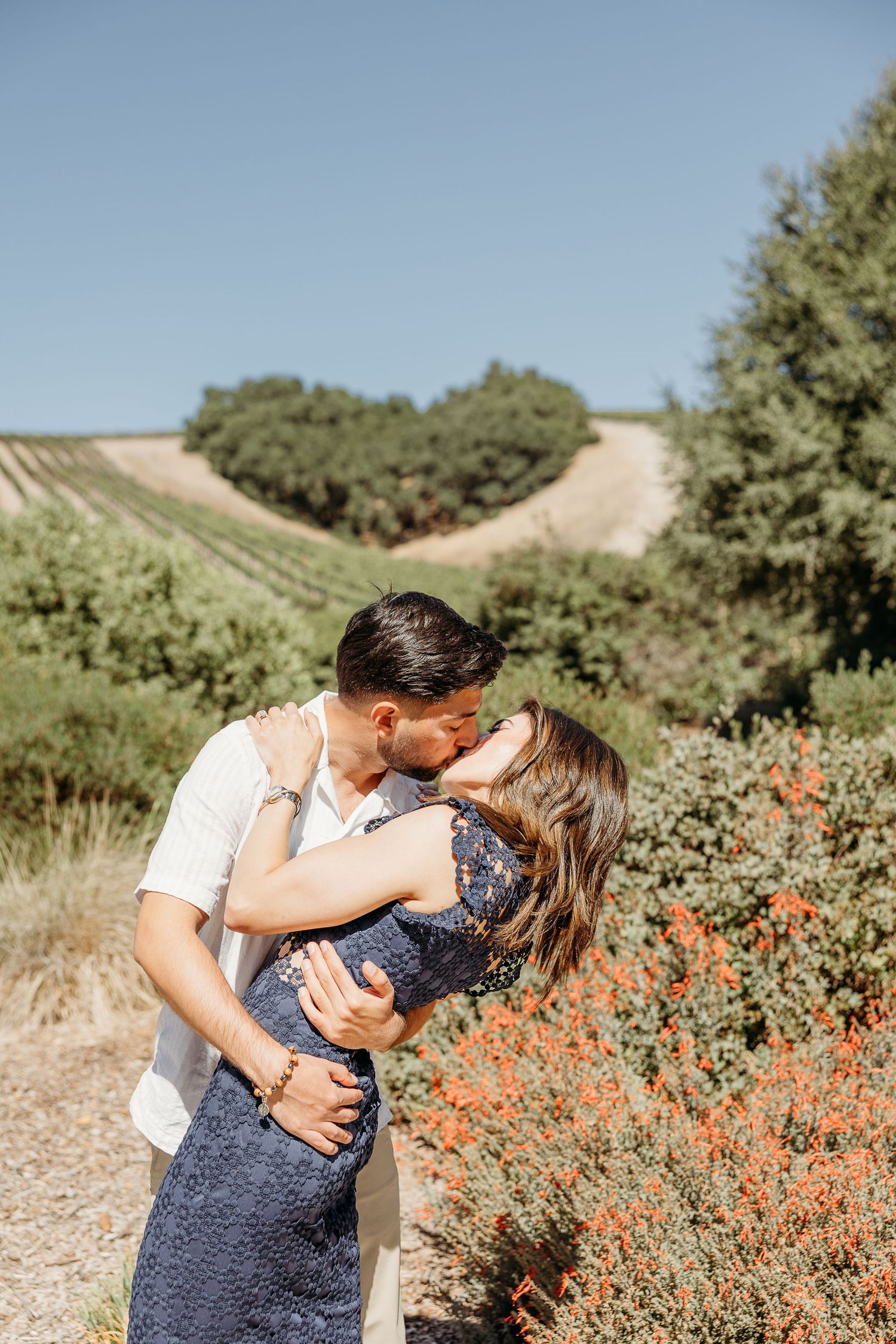 Romantic embrace captured in sunny meadow with blooming orange flowers and rolling hills.