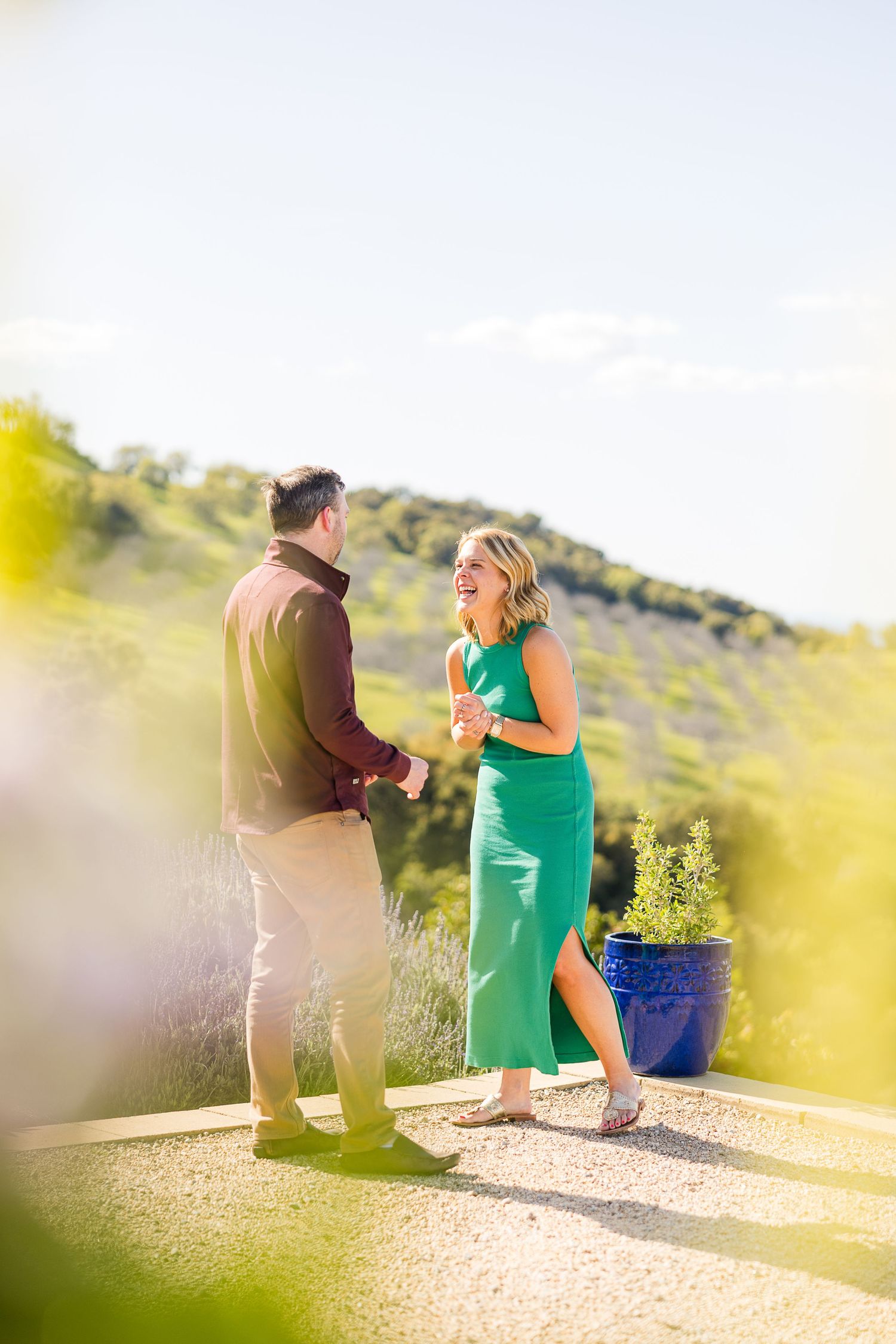 A couple celebrates their engagement on a sunny hillside path with a blue planter in the background.
