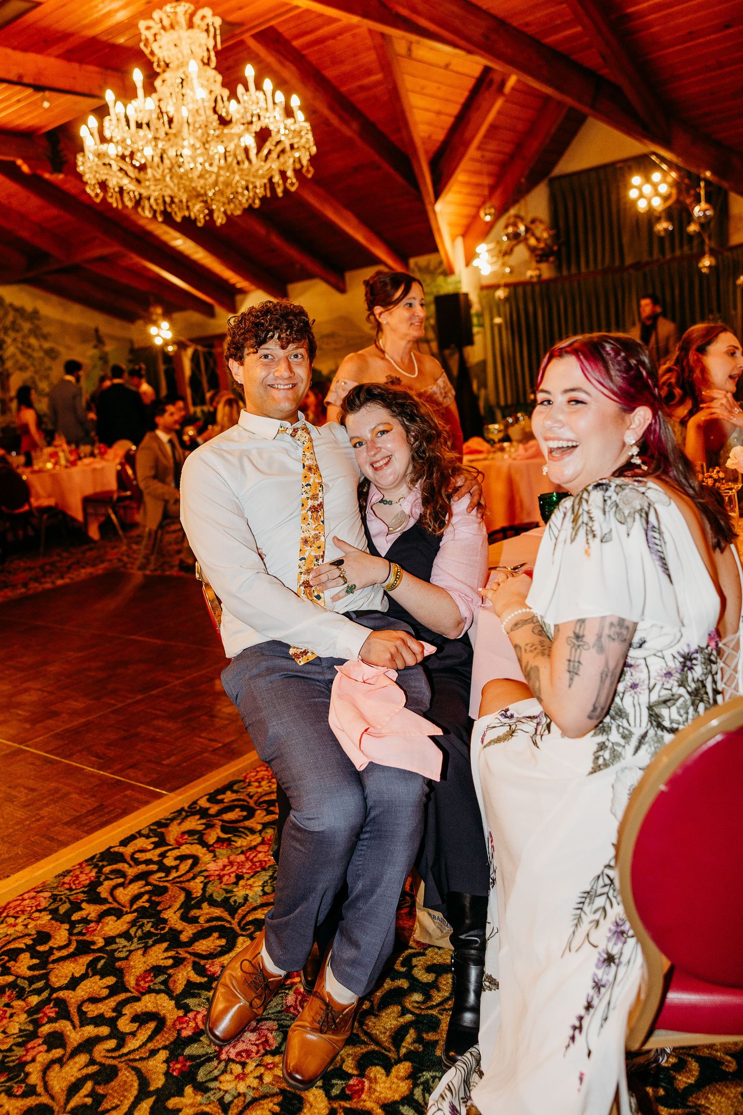 Wedding guests dance and celebrate under warm chandelier lighting in a rustic reception venue with wooden beams.