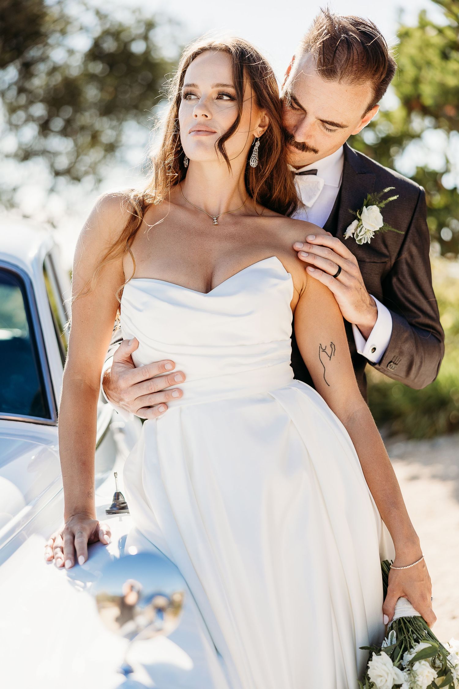 Elegant wedding couple poses against classic car in sunlit outdoor setting.