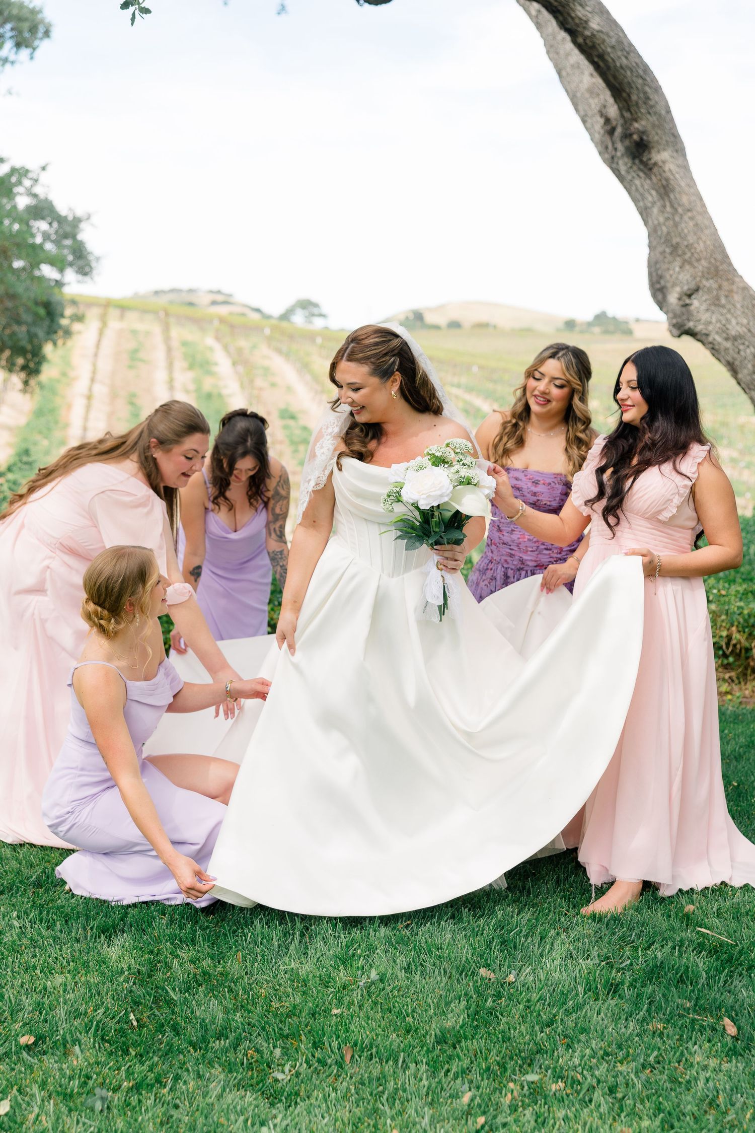 Bridesmaids in pink and lavender gowns helping bride with white dress on vineyard lawn.