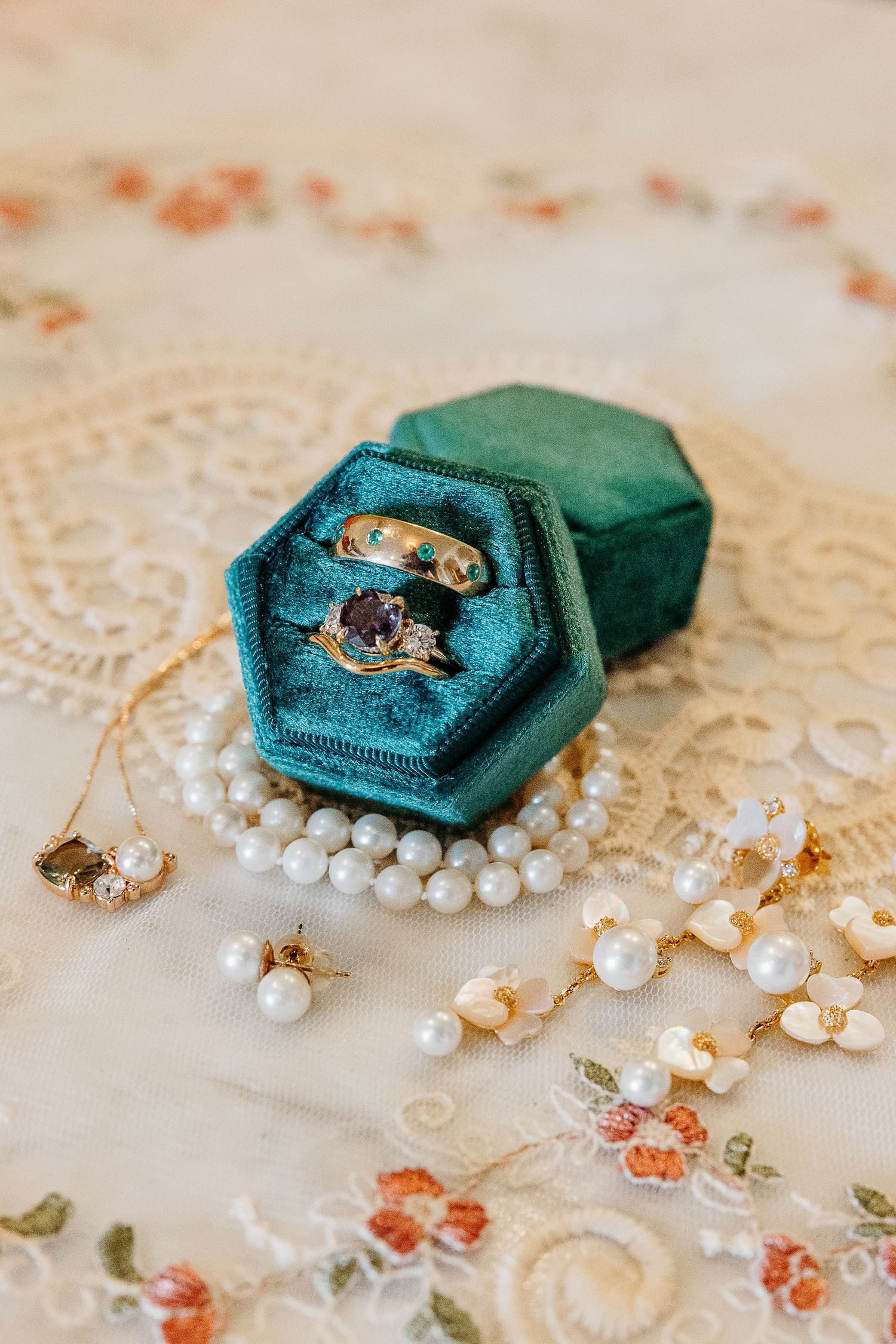 Wedding rings displayed on a teal velvet hexagonal ring box with delicate pearl jewelry accents.