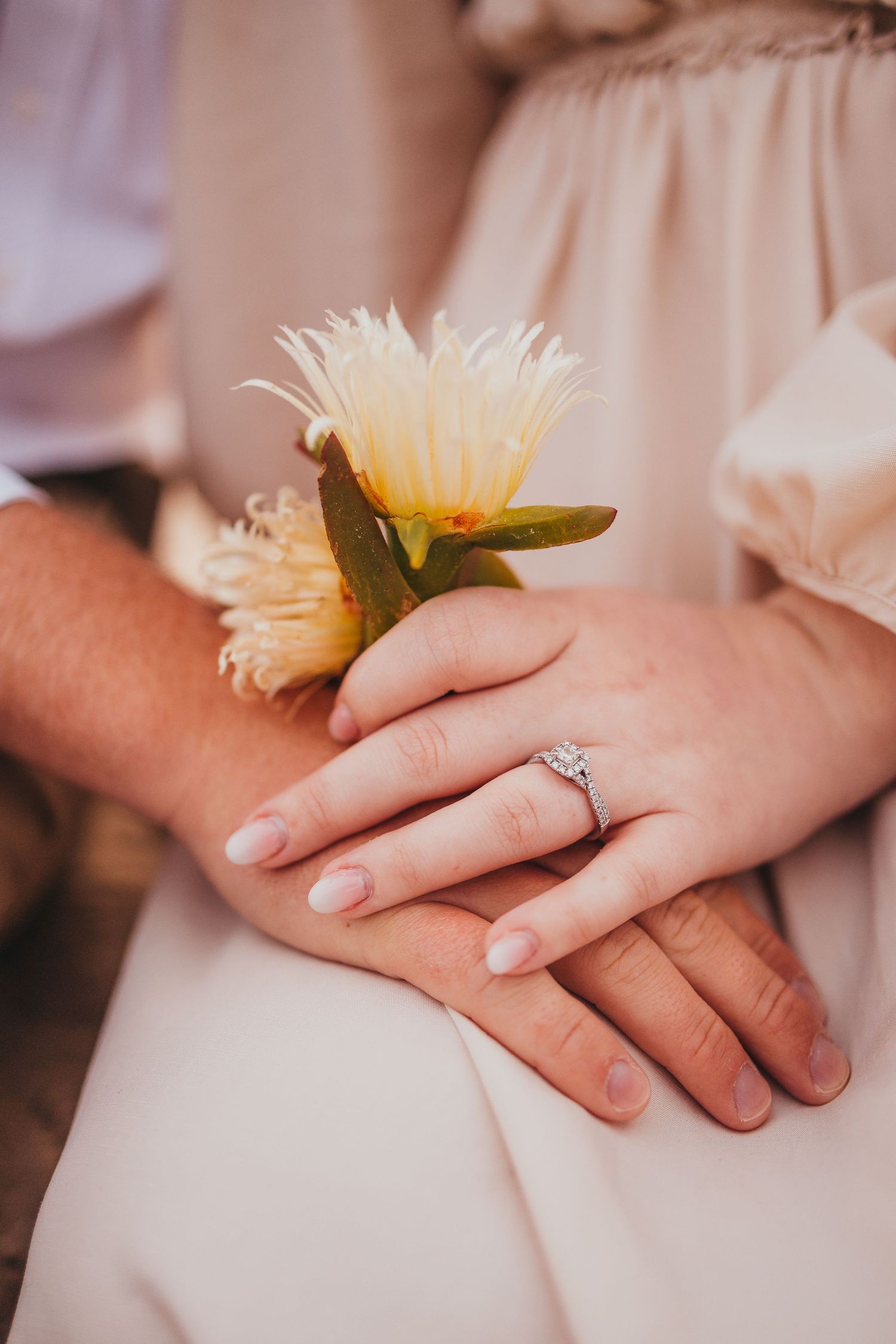 Detail shot of hands with engagement ring and yellow flower on light fabric.