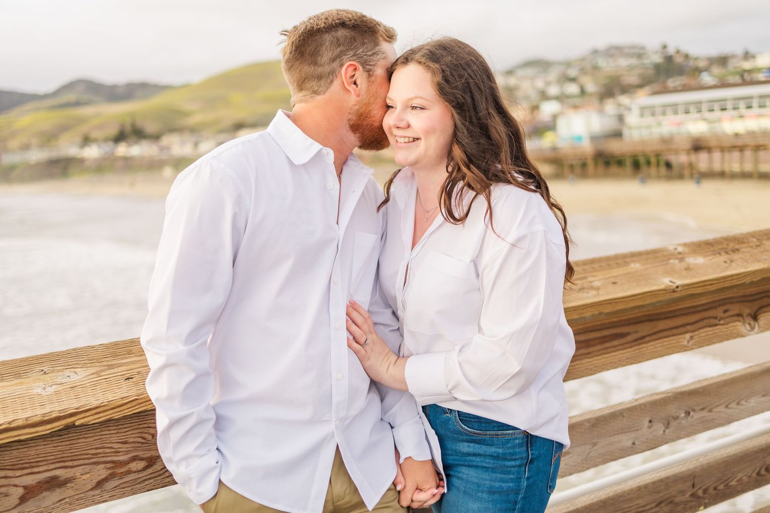 A couple in white shirts embracing on wooden pier with coastal scenery.