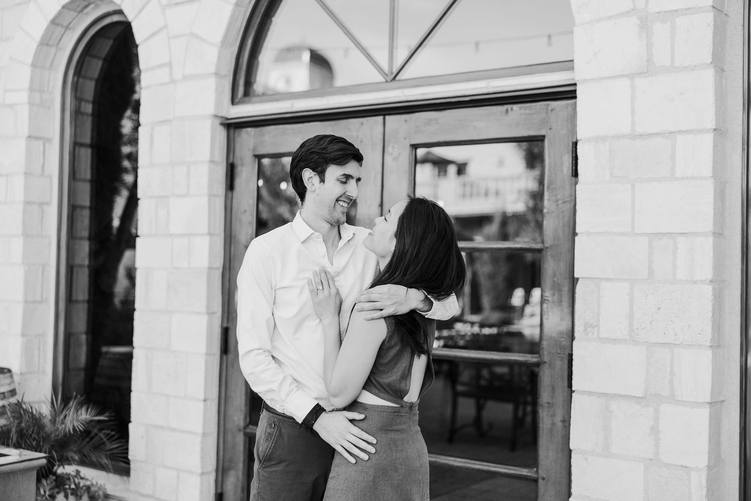 A black and white photo of a couple sharing an intimate moment by arched windows.