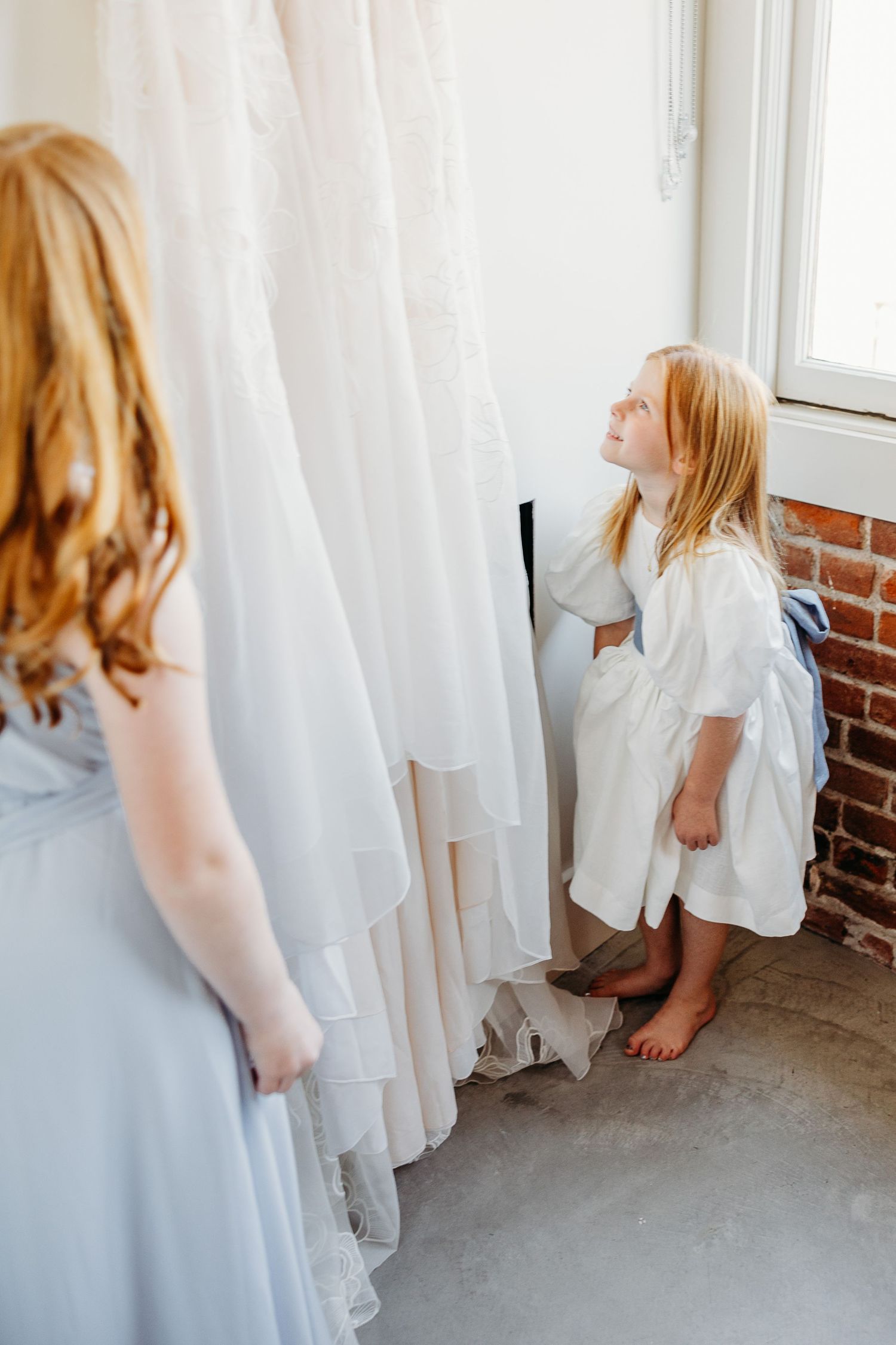 A young flowergirl in white examines wedding dresses hanging on a wall.
