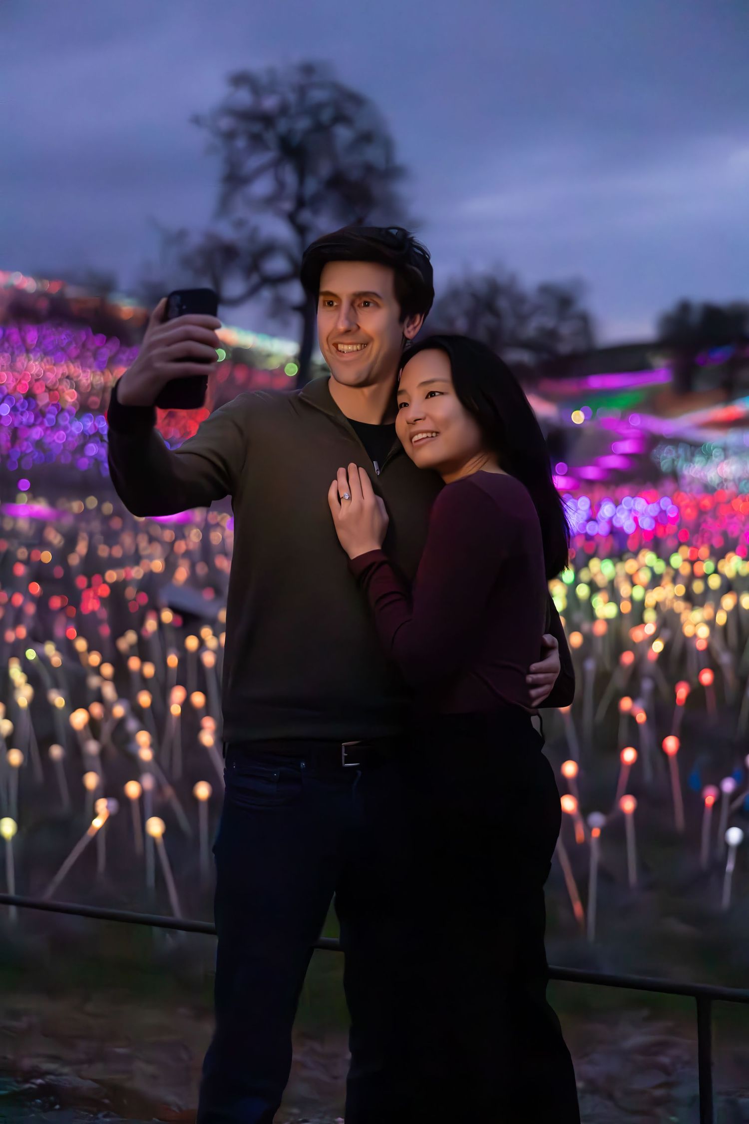 A couple taking selfies together at dusk with colorful illuminated light displays in the background.