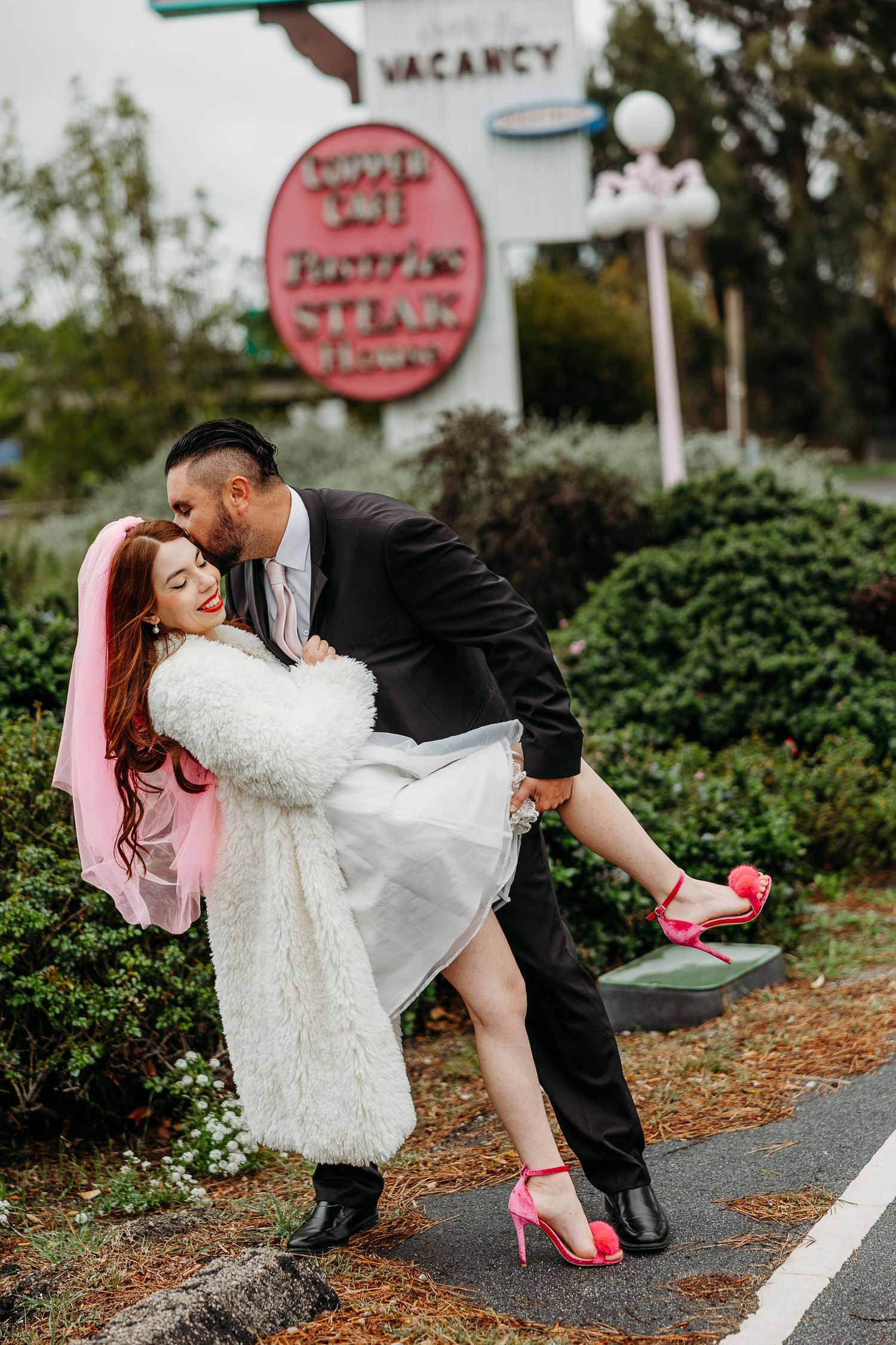 Romantic couple share an intimate moment in front of a vintage-style neon sign wearing formal attire and bright pink shoes.