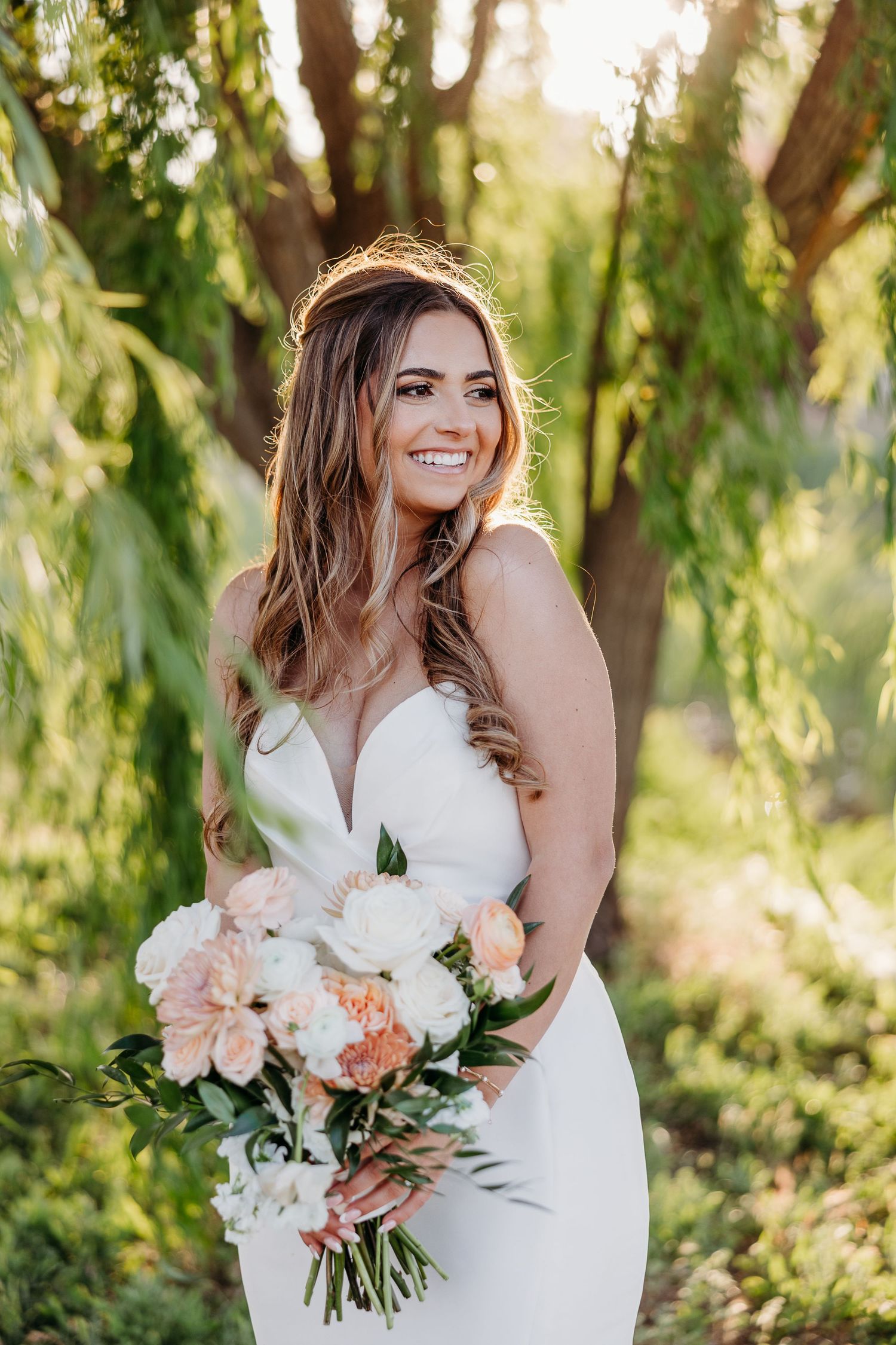 A bride in a white wedding dress holds a beautiful peach and white bouquet against a backdrop of willow trees.
