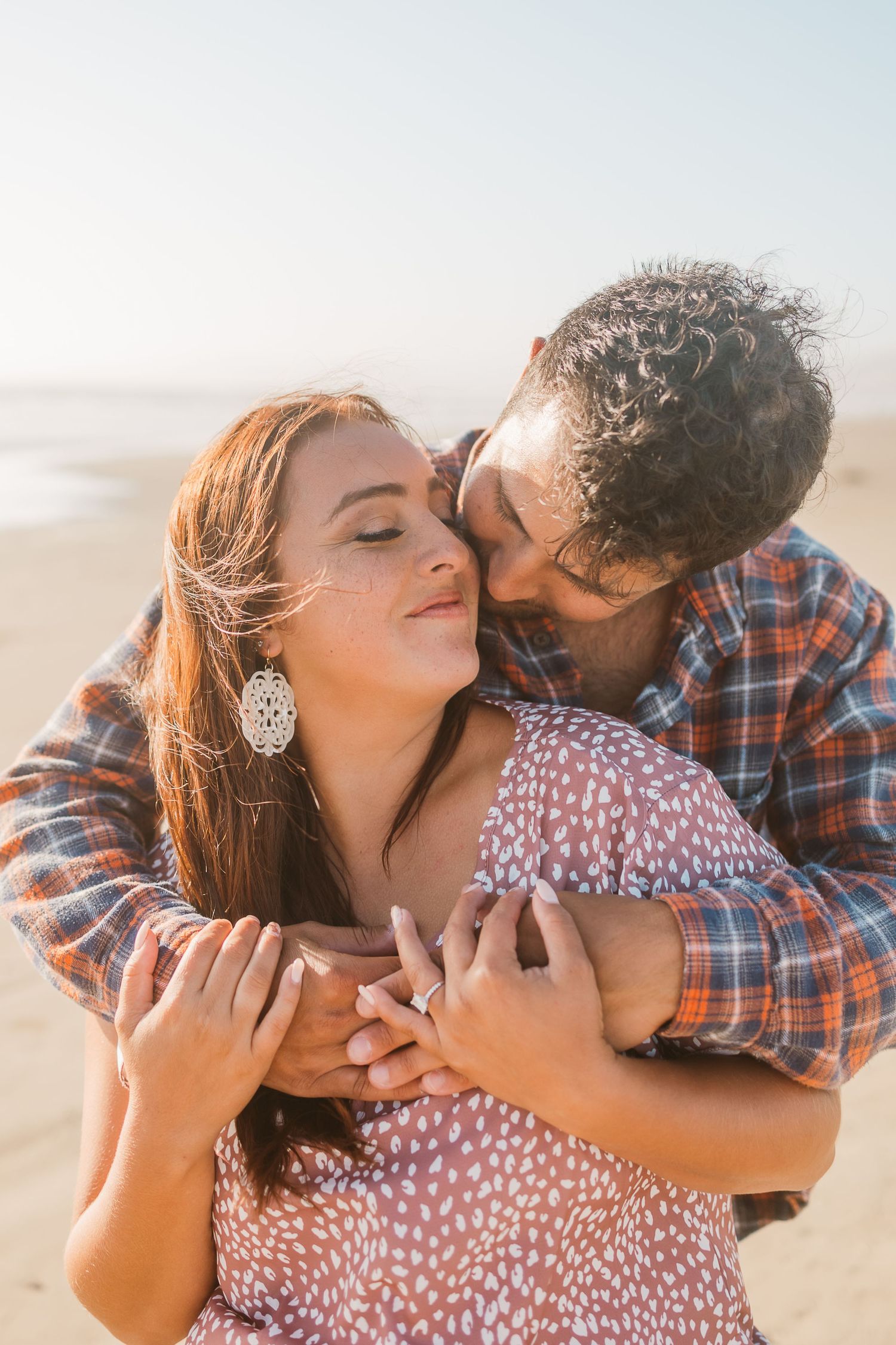 Romantic couple embracing on beach at sunset with woman in polka dot dress and man in plaid shirt.