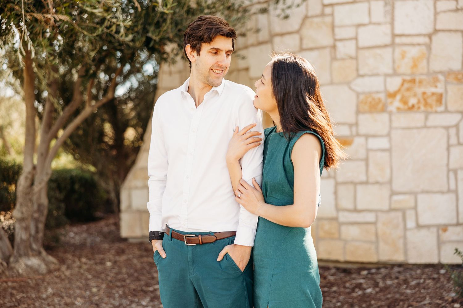 A couple in coordinating green and white outfits share a romantic moment in front of a stone wall.