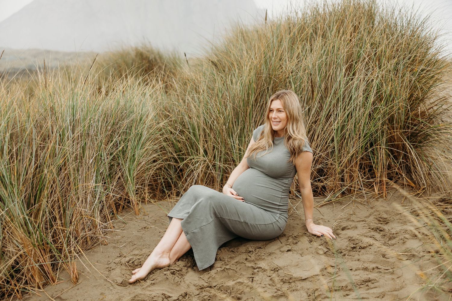 Maternity photo of expectant mother in gray dress reclining on sandy beach near grass.