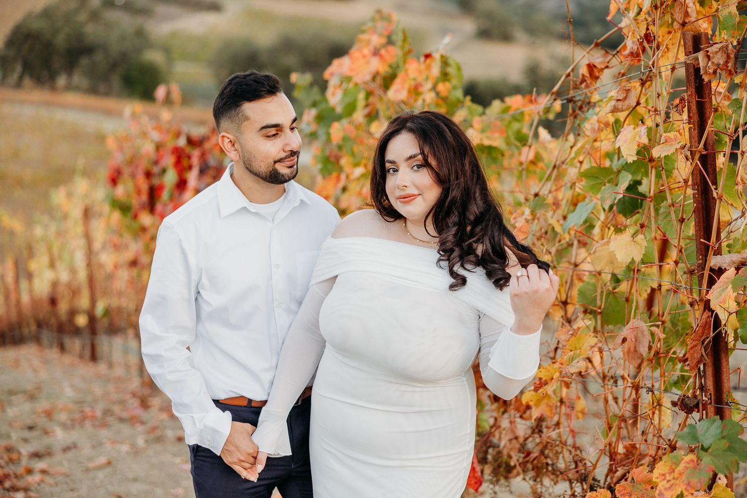 Couple poses in autumn vineyard with orange and red fall foliage during sunset.
