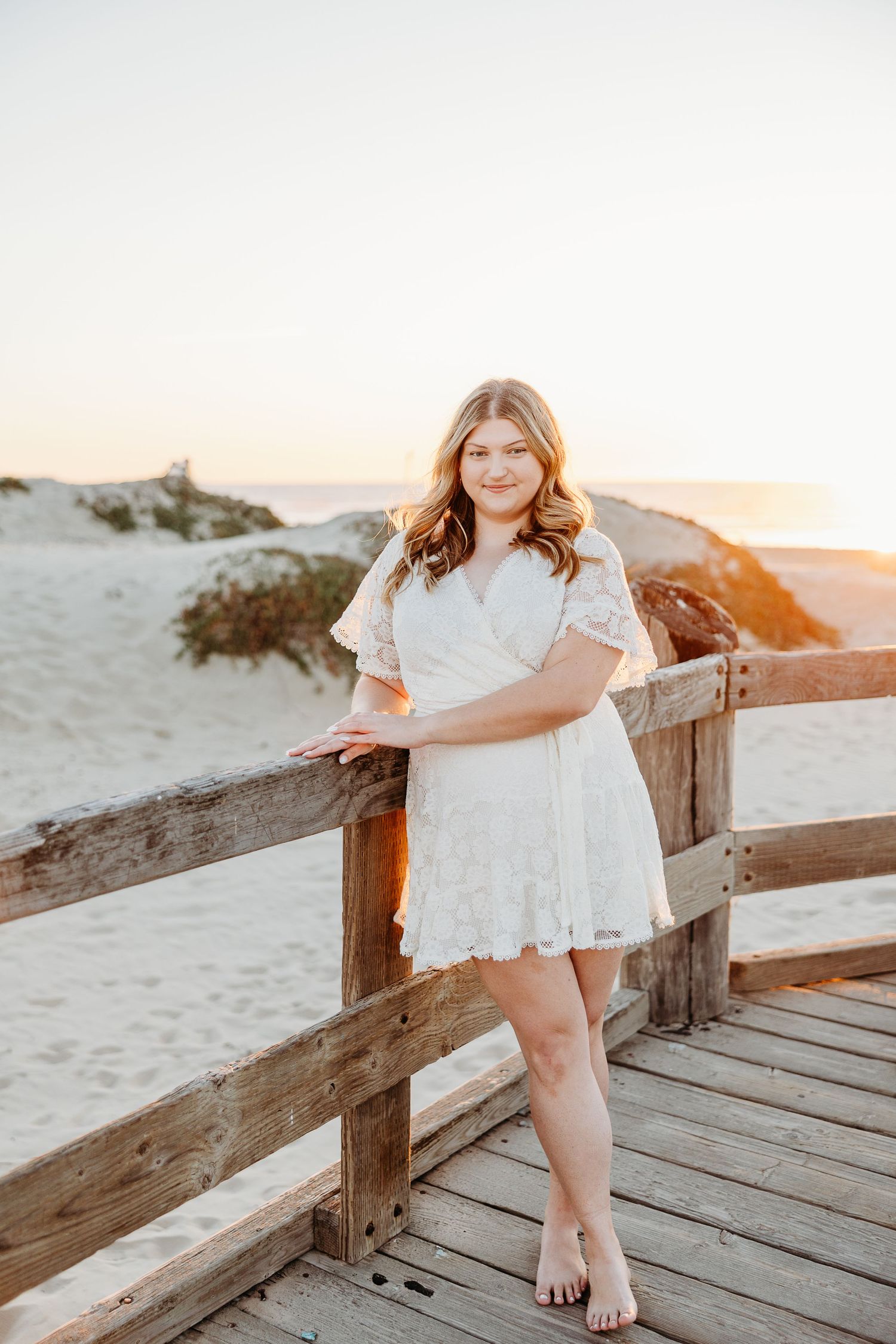 Someone in a white dress poses barefoot on a wooden boardwalk at the beach during sunset.