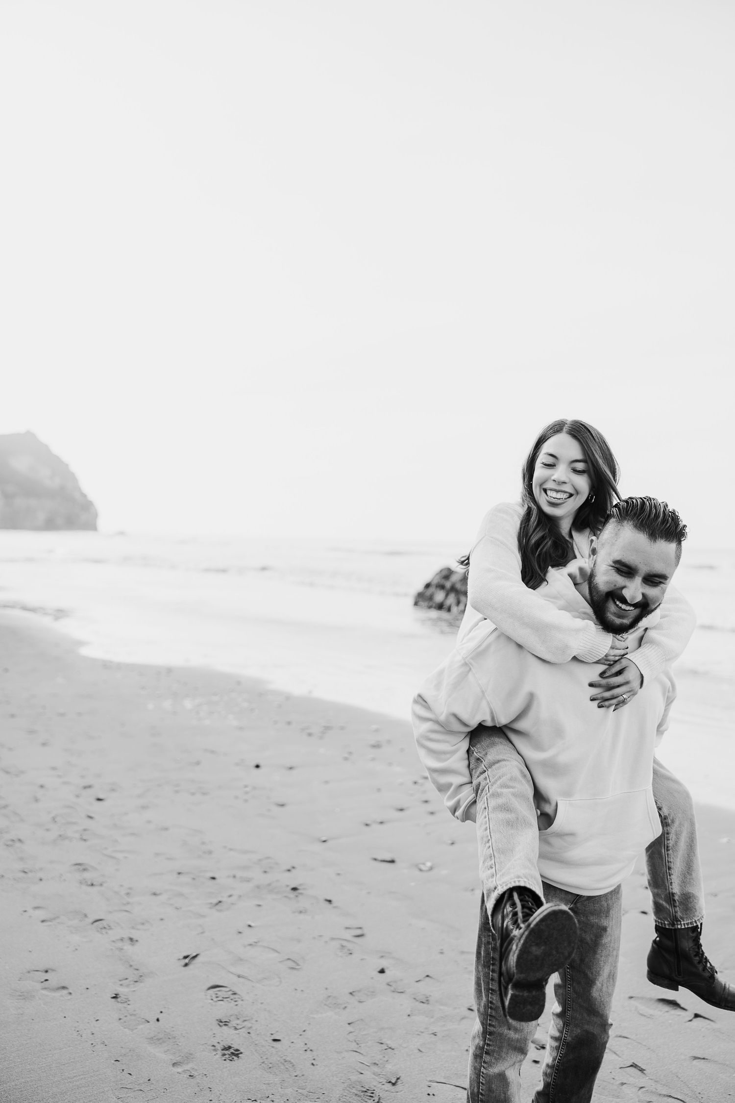 A joyful piggyback ride moment captured in black and white on a scenic beach with coastal cliffs in the background.