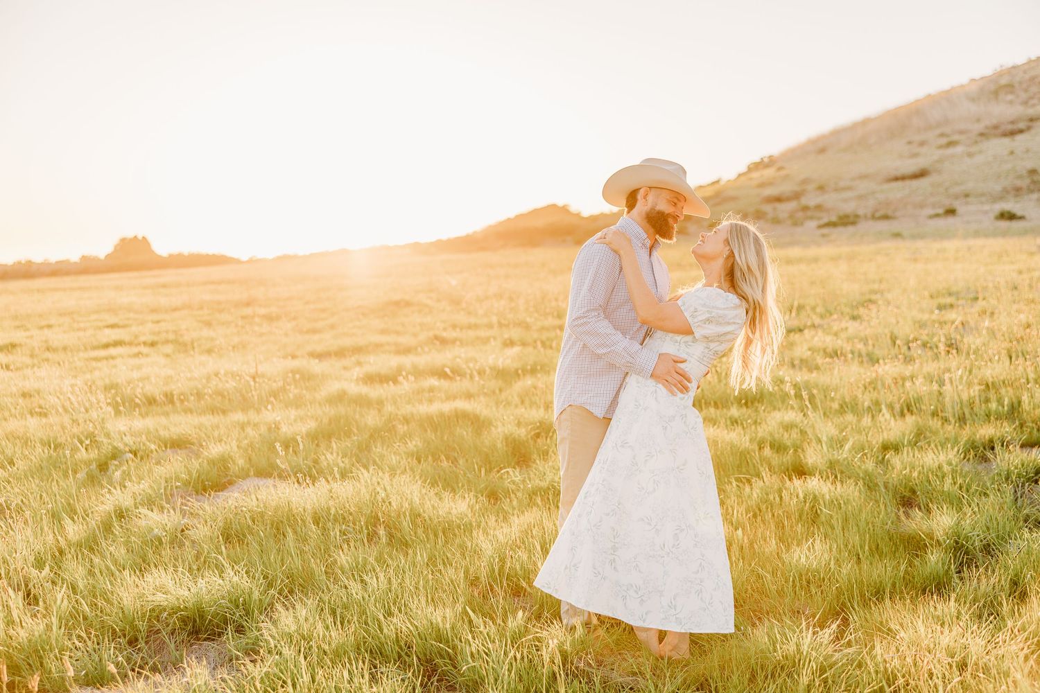 A couple embraces in a golden sunlit meadow wearing light summer attire.