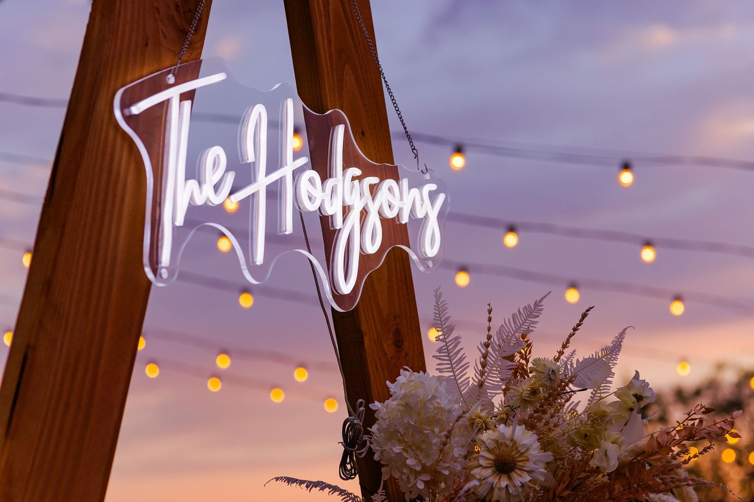 Neon sign and dried floral arrangement against wooden A-frame structure at dusk.