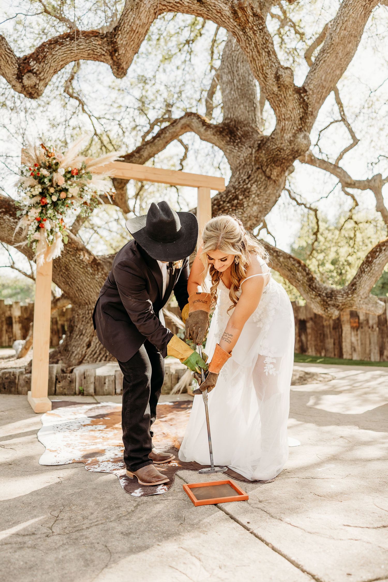 A couple in western wedding attire lights a unity candle together under a rustic wooden arch and oak tree.