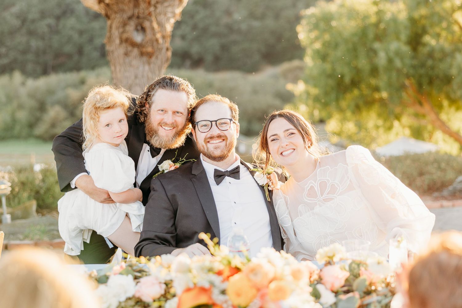 Wedding party celebrates at an outdoor reception table with floral decorations at sunset.