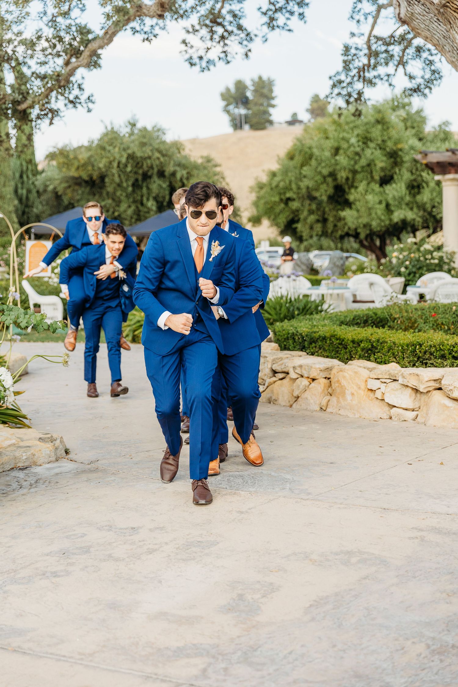 Group in navy blue suits walking along a garden path during a wedding celebration.
