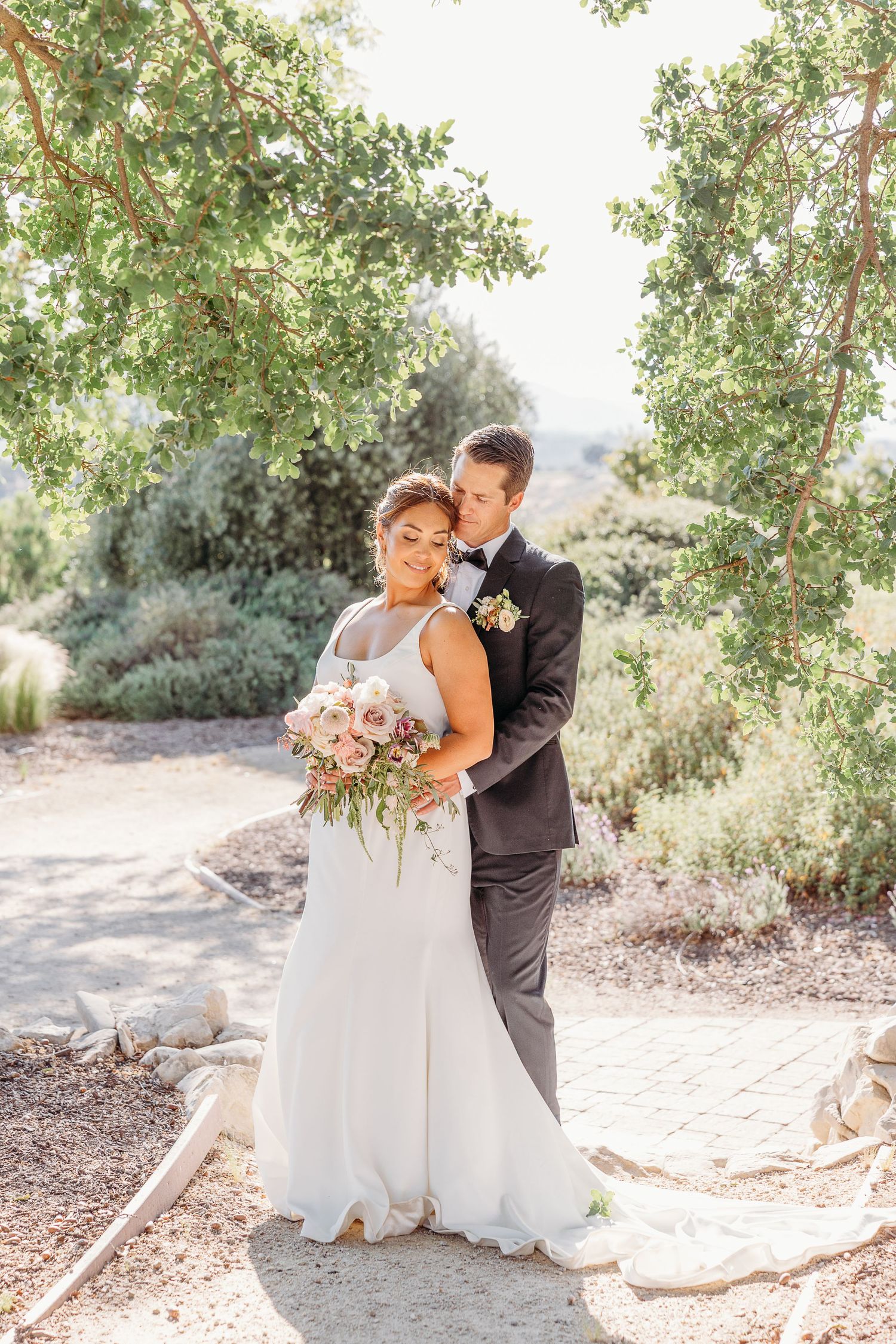 A romantic wedding couple embraces outdoors under sunlit trees in a natural garden setting.