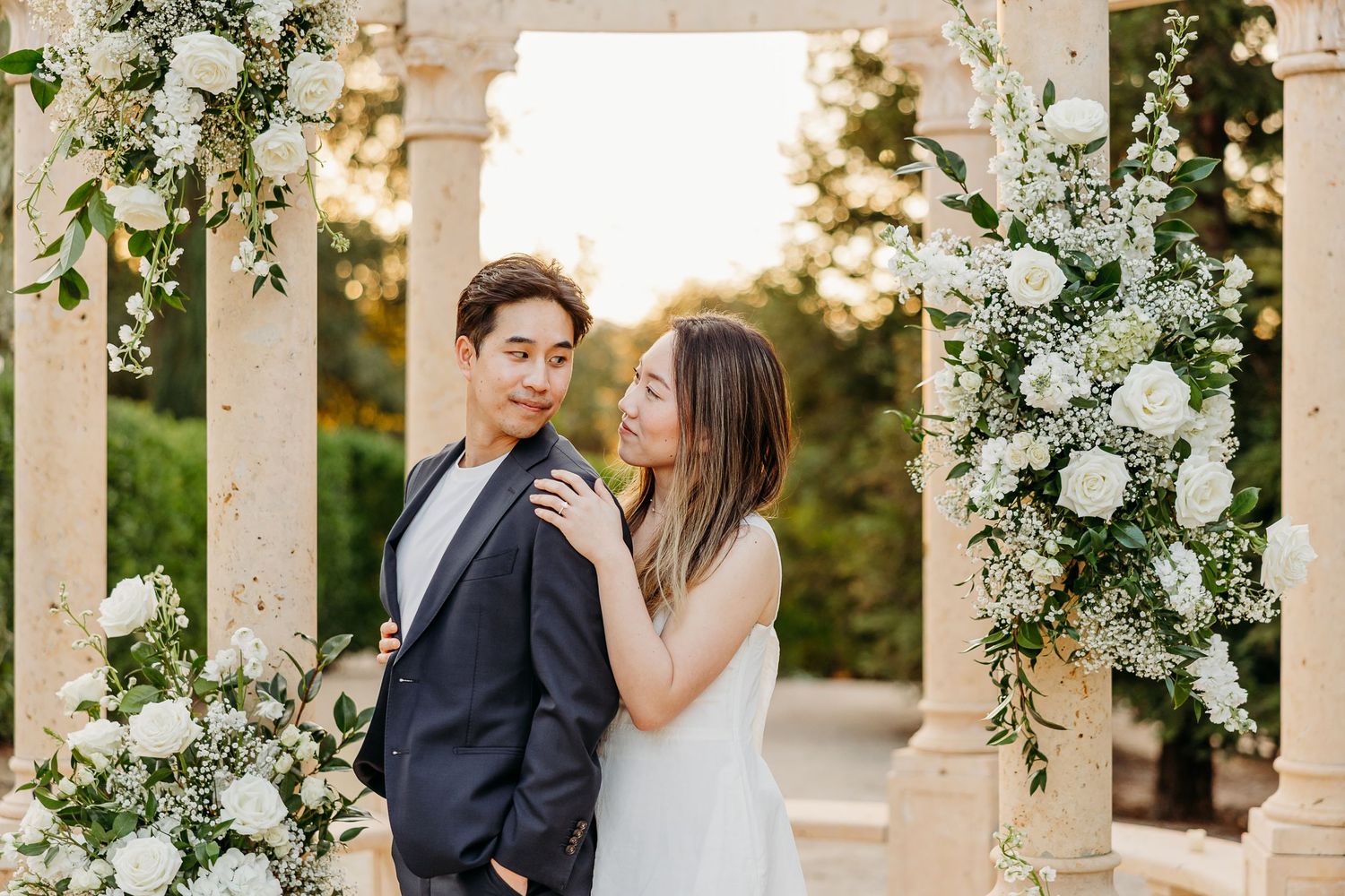 Elegant wedding portraits under a white floral arch with greenery and roses.