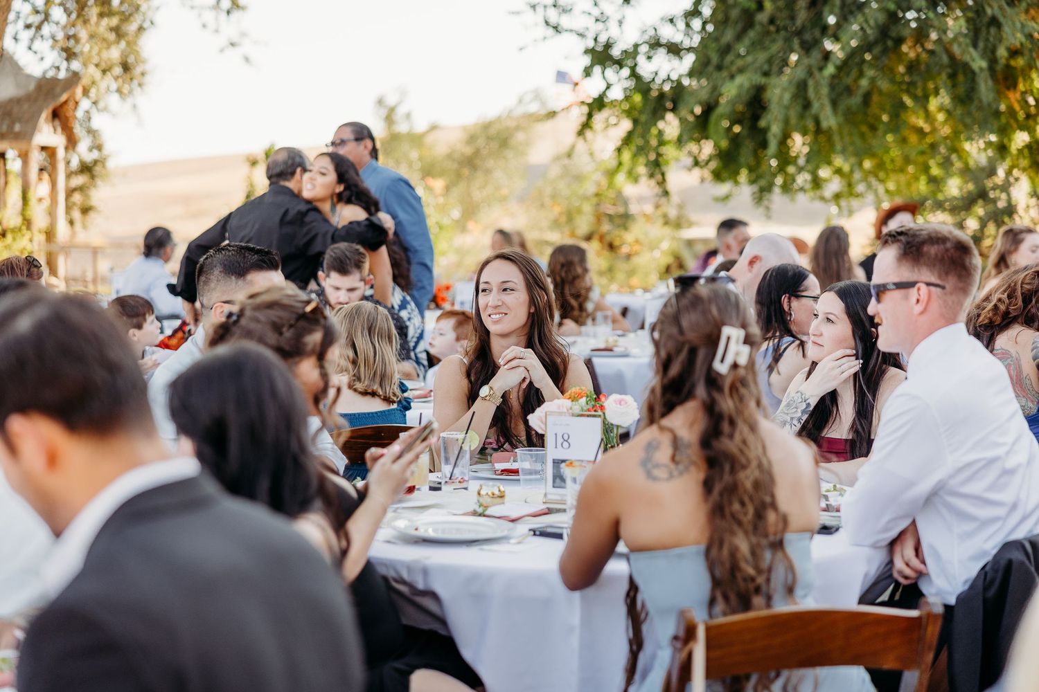 Large group of wedding guests dancing and celebrating at an outdoor reception under trees.