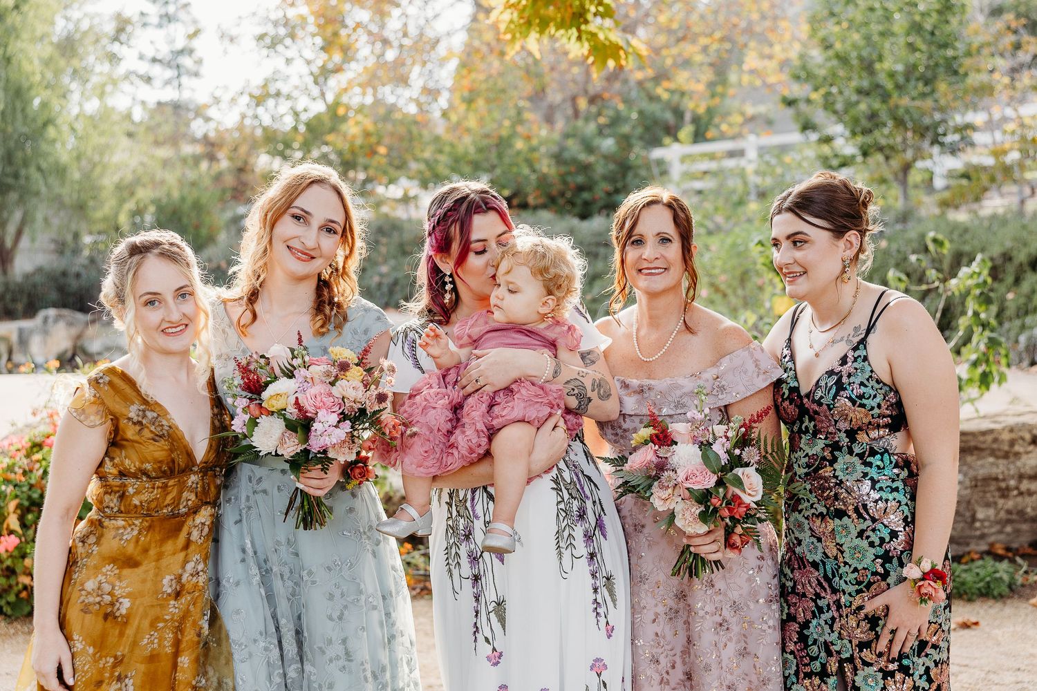 A wedding party wearing floral dresses poses together in an outdoor garden setting during autumn.