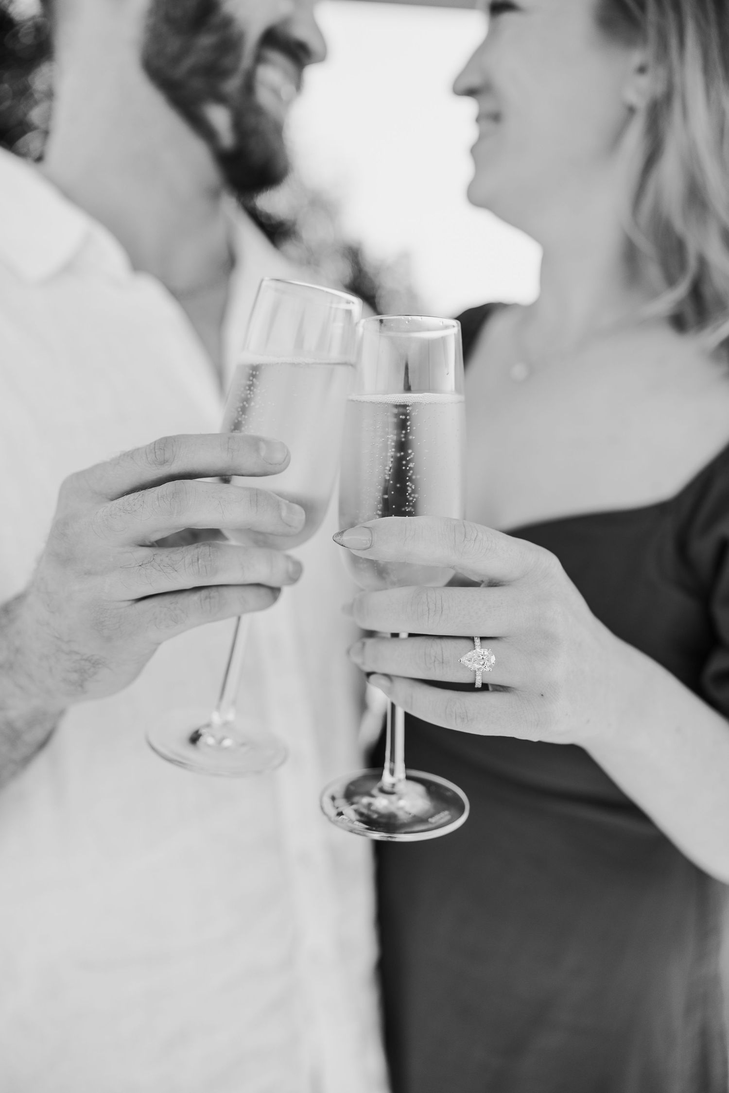 Black and white photo of couple toasting with champagne flutes, engagement ring visible on woman's hand.
