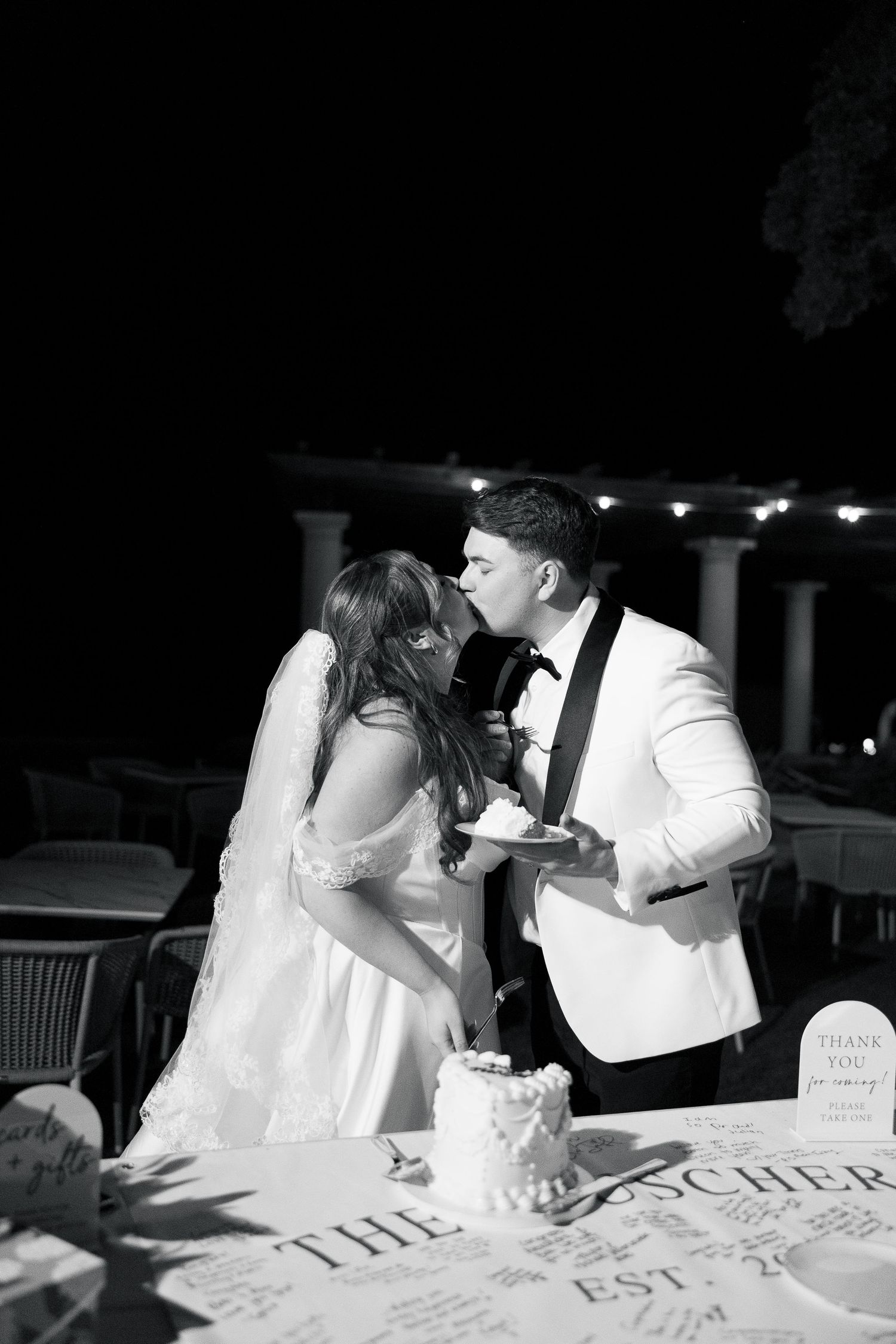 Bride and groom share a kiss while cutting wedding cake at elegant outdoor evening reception in black and white.
