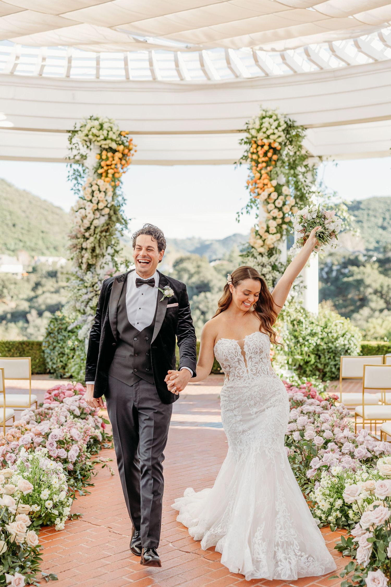 Joyful wedding couple dancing and celebrating under a white pergola decorated with colorful floral arrangements.