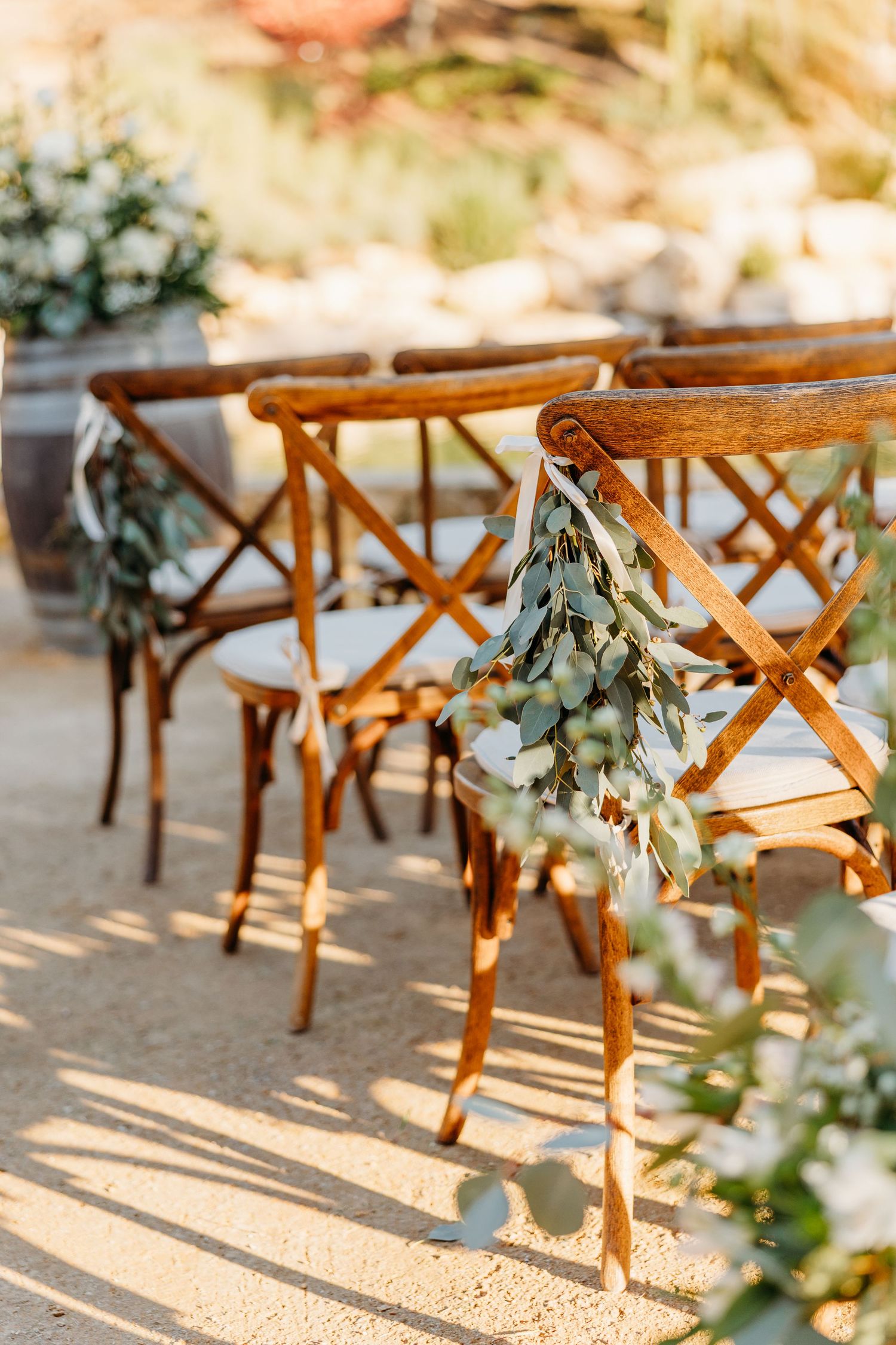 Wooden cross-back chairs decorated with floral arrangements line up for an outdoor wedding ceremony.