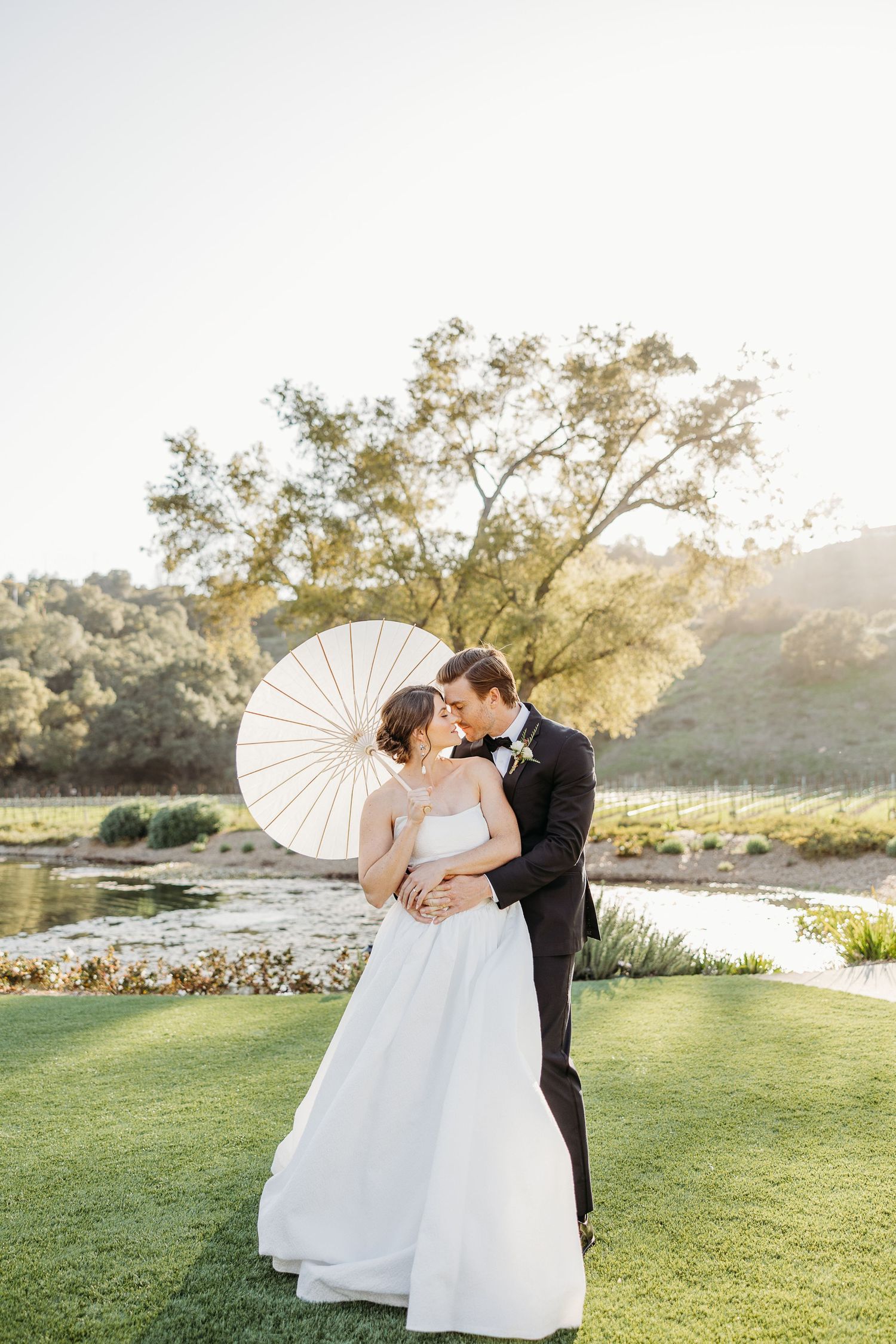 Wedding couple with parasol shares romantic moment on sunlit garden lawn.