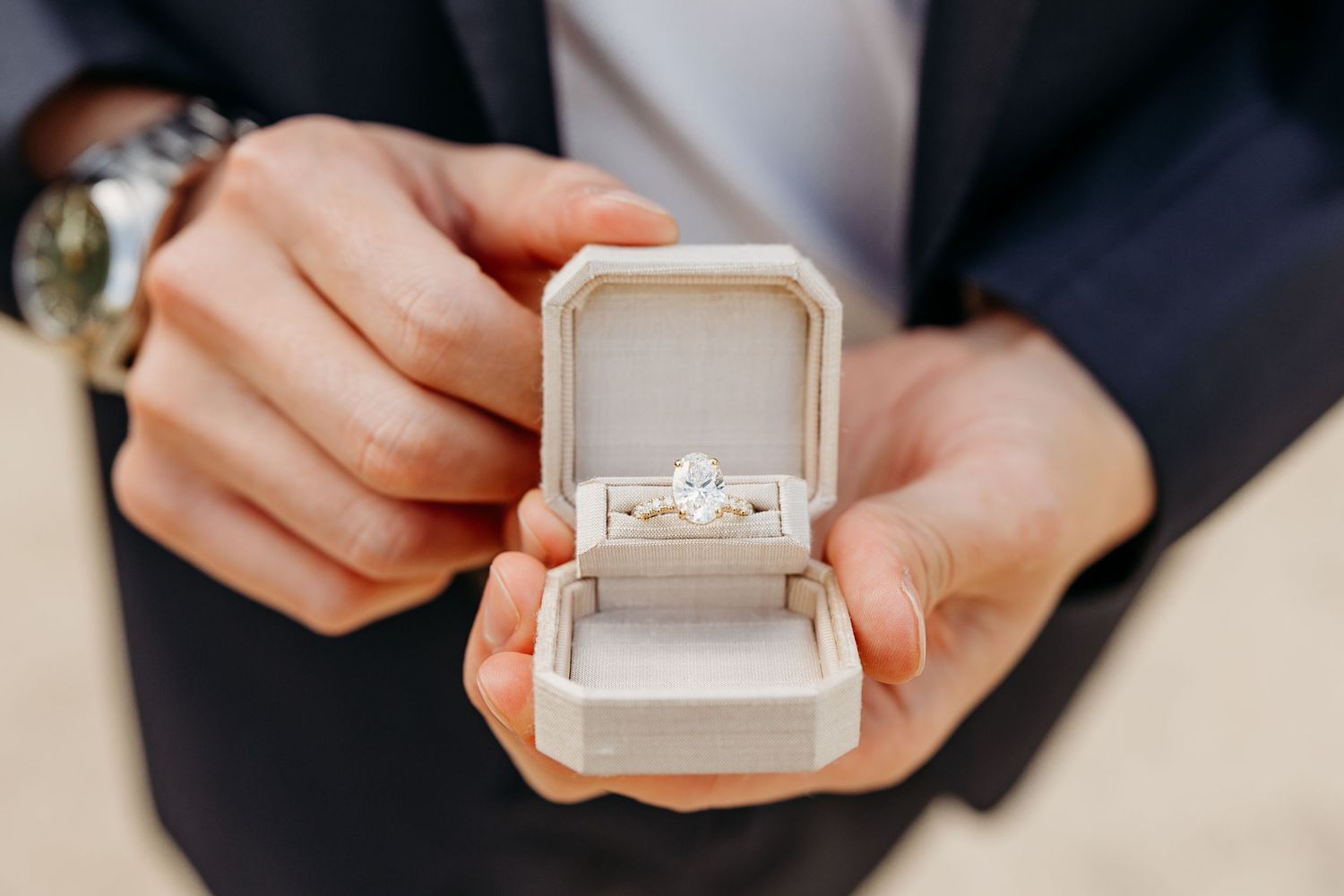 Close-up view of hands holding a light gray velvet ring box in a wedding ceremony setting before a surprise proposal