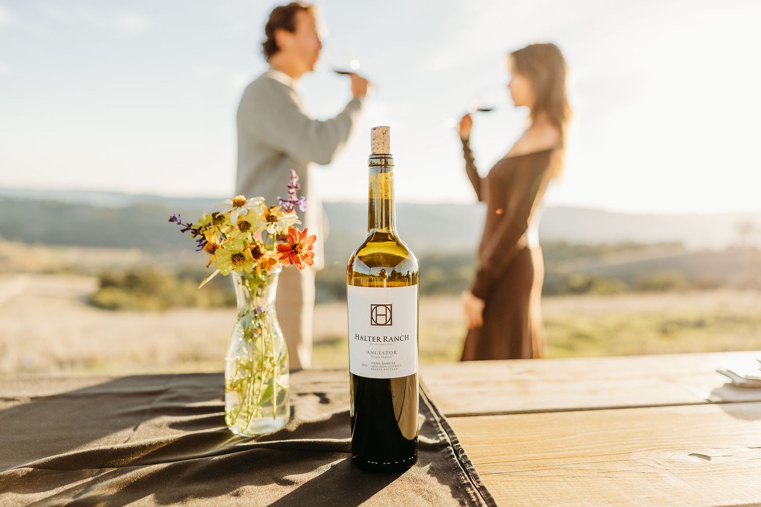 Wine bottle and glasses on wooden table at sunset with mountain views.