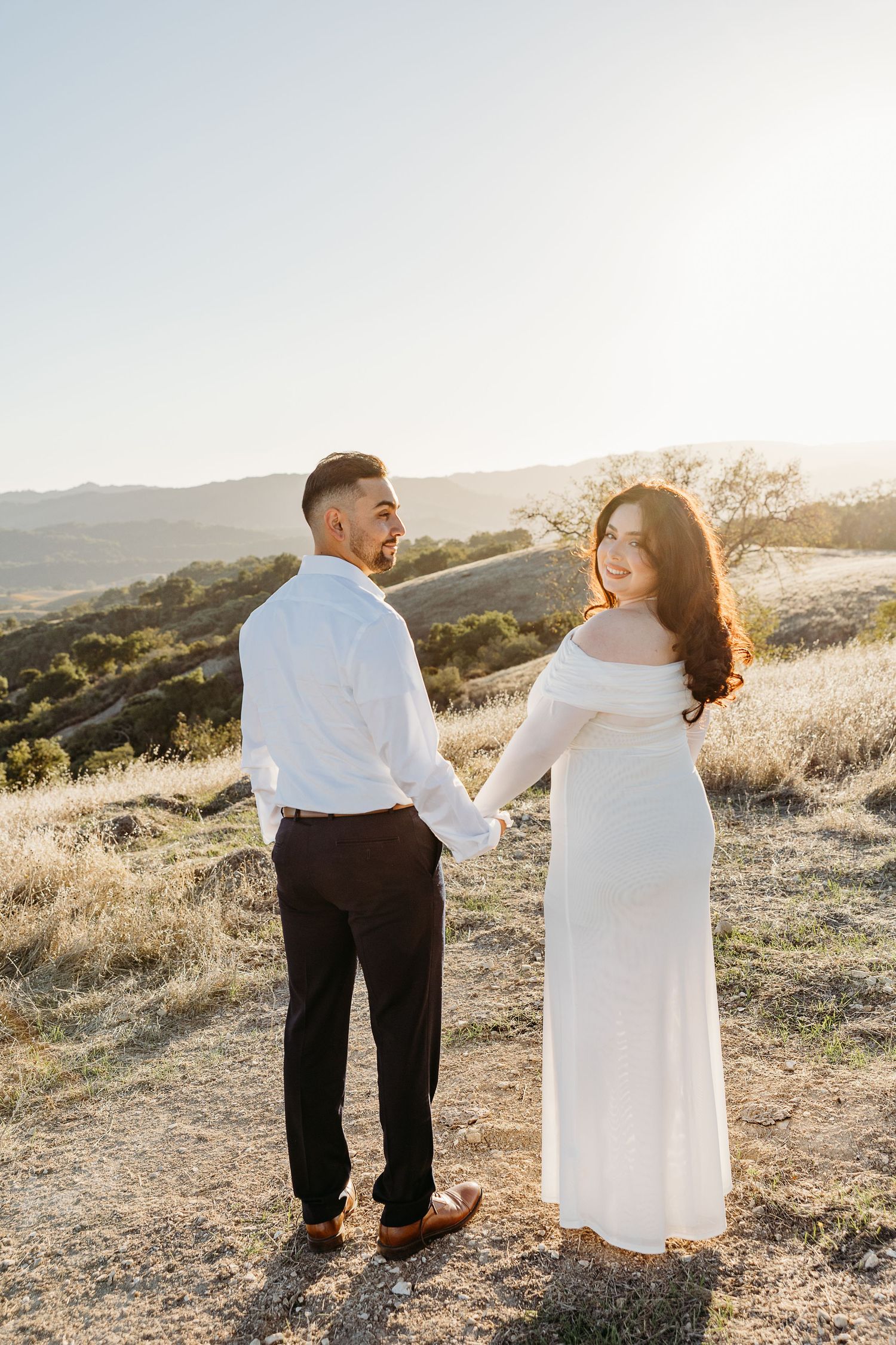 A couple in formal attire stands together on a hilltop during golden hour with mountains in the background.
