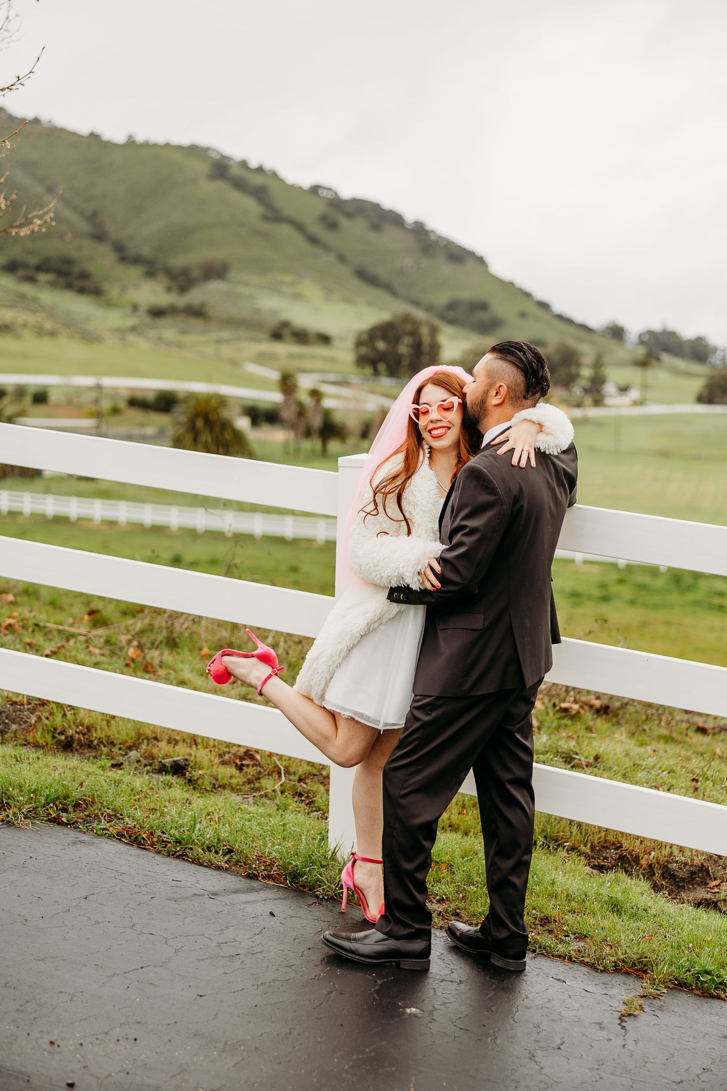 A couple shares a romantic moment by a white fence on a rainy day with green rolling hills in the background.